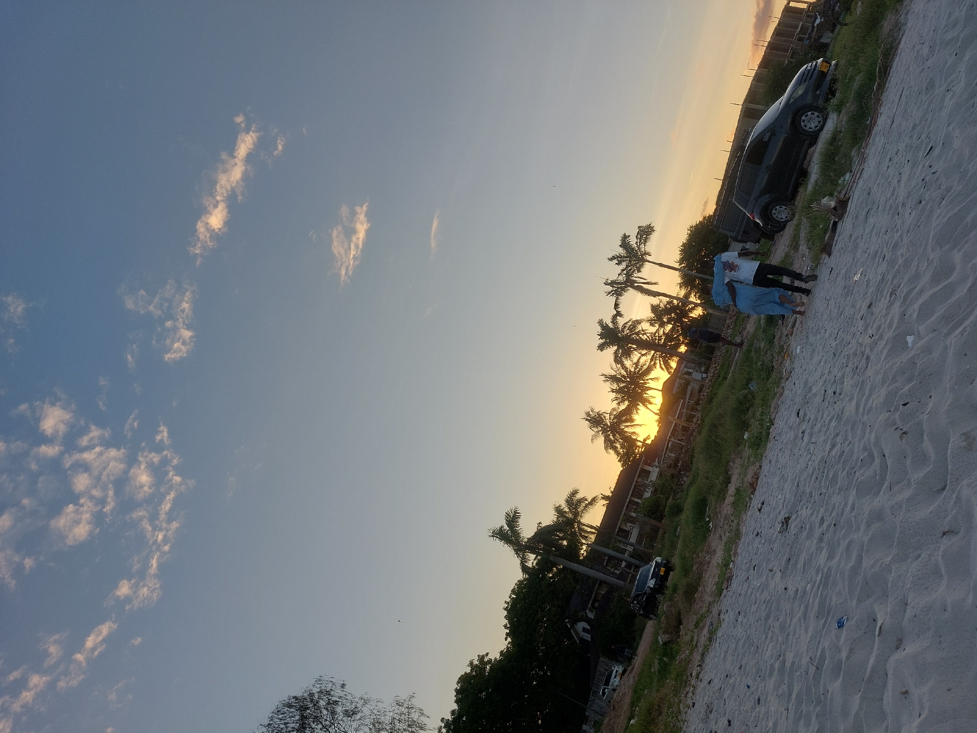 The image depicts a beach scene at sunset. The sky is a gradient of blue to orange, with scattered clouds. Silhouetted palm trees line the horizon, partially obscuring the setting sun. Two people, possibly a couple, are walking along the sandy beach. A dark-colored vehicle is parked nearby. The scene is likely located along the coast of Dar es-Salaam, Tanzania, given the presence of palm trees and the beach environment. There are some scattered debris on the beach.