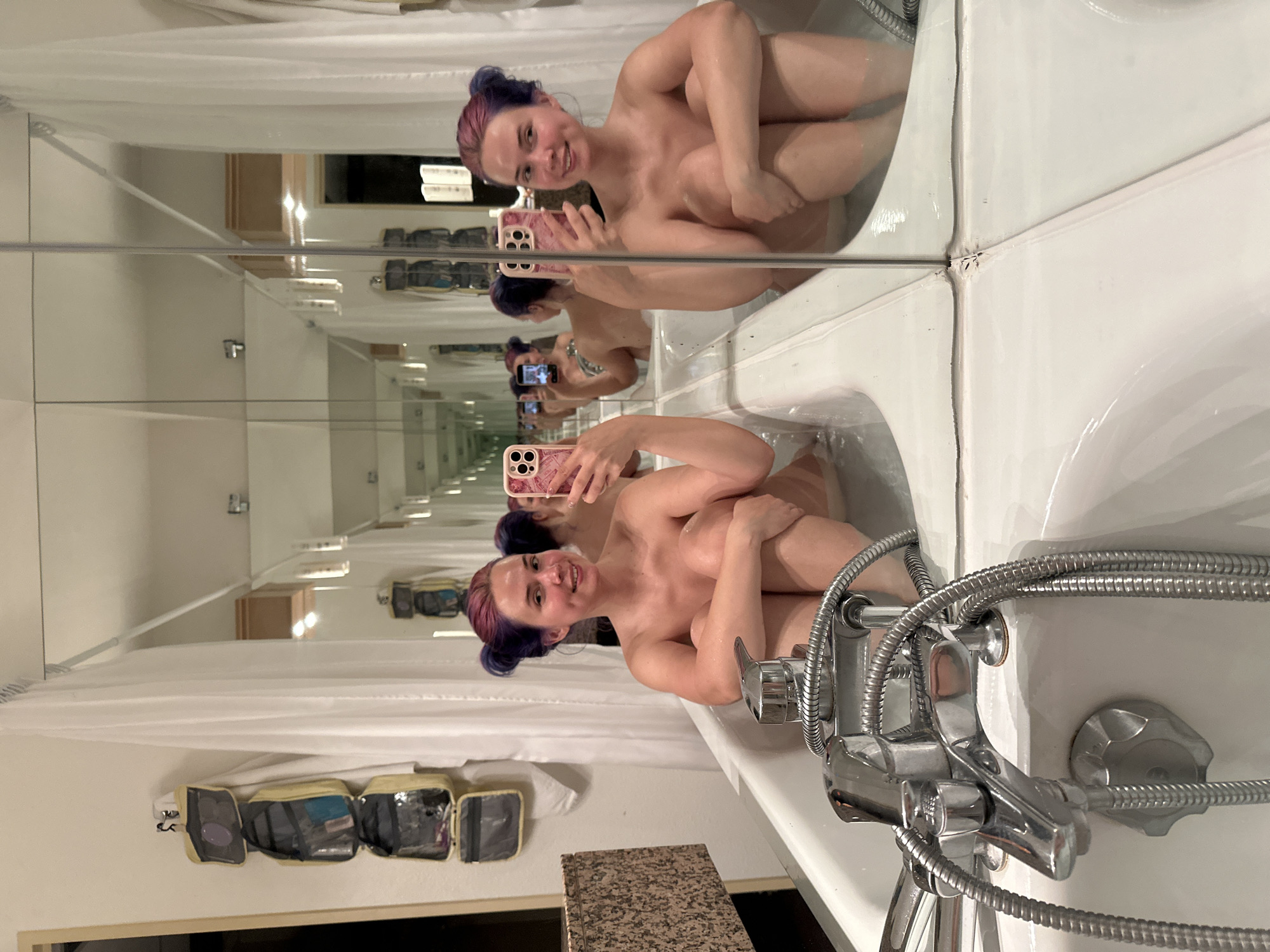 A nude woman with purple hair is sitting in a bathtub, holding a phone and taking a selfie. The bathtub is white and has a silver faucet. The bathroom is tiled in white. There is a mirror above the bathtub, which creates an infinity effect. The location appears to be a hotel bathroom in Ramonville-Saint-Agne, France. The wall to the left of the bathtub has a rack of hanging bags.