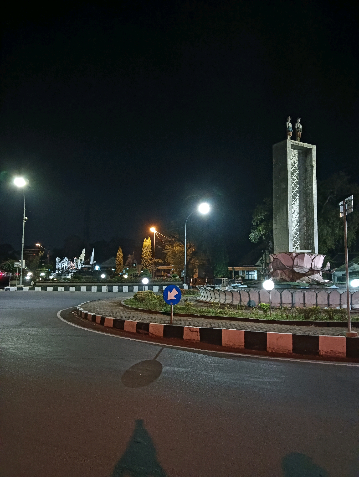 The image depicts a roundabout at night in Purworejo, Indonesia, specifically the Tugu Pahlawan or Heroes' Monument roundabout. The roundabout features a tall, rectangular monument topped with two statues of men in uniform. At the base of the monument is a decorative fountain resembling a lotus flower. Several streetlights illuminate the scene, casting shadows on the wet asphalt road. A blue directional sign with a white arrow pointing left is visible in the foreground. The roundabout itself is bordered by a curb with alternating black and white segments, and landscaped with low-lying plants. In the background, another roundabout with a large sculptural display, possibly depicting a historical scene with horses and figures, is illuminated by lights. Trees and buildings are also visible in the background, shrouded in the night darkness. The overall atmosphere is serene, with the illuminated monuments and roundabout creating a focal point in the otherwise dark urban environment.