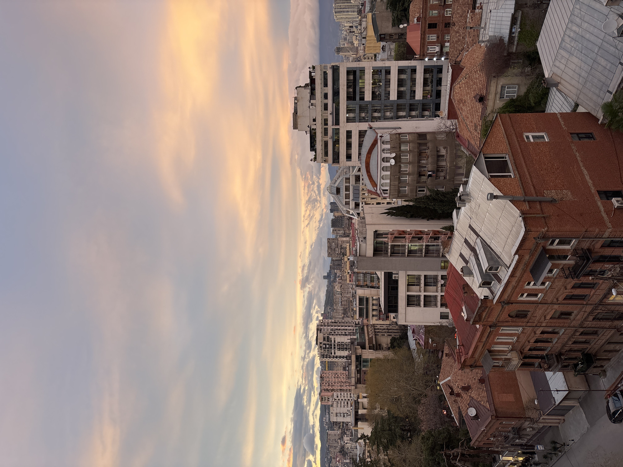 The image is an elevated, slightly rotated view of the city of Tbilisi, Georgia, during either sunset or sunrise, suggested by the warm, pastel-colored sky with striated clouds. The urban landscape features a dense mix of architectural styles, including modern high-rise buildings and older, traditional brick structures with sloped, tiled roofs. The buildings are interspersed with trees and greenery. The cityscape extends into the distance, blurring slightly towards the horizon. The foreground is dominated by the rooftops of closer buildings, offering a detailed view of their materials and designs.