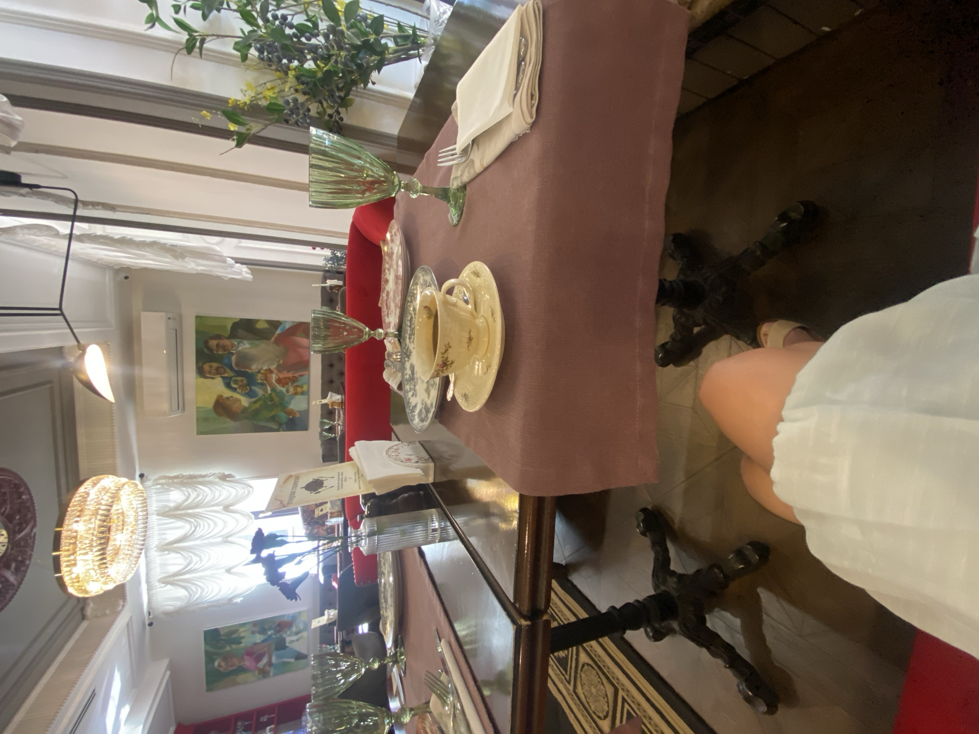The image shows a table setting in a restaurant. The table is covered with a mauve tablecloth and set with plates, glasses, and cutlery. A teacup with a saucer is placed in the center of the table, filled with a dark liquid, likely coffee or tea. Behind the teacup, there is a vase of flowers, and further in the background, a painting hangs on the wall. The restaurant appears to be well-decorated, with a chandelier hanging from the ceiling. The lighting suggests it might be daytime or early evening. The lower part of the image shows a person's legs, suggesting they are seated at the table. The location is identified as Tomsk, Russia.