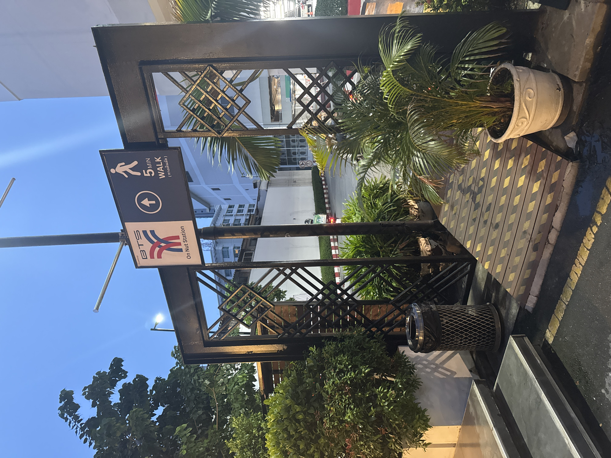The image shows an exterior shot at dusk or dawn, possibly in Bangkok, Thailand, as indicated by the presence of a BTS (Bangkok Mass Transit System) sign. The sign, mounted on a black metal structure, reads "BTS On Nut Station" and also displays a directional arrow with "5 MIN WALK". The overall scene depicts an entrance or pathway leading to the station.
In the foreground, to the left, there are two steps leading up to a walkway. Next to the steps is a metallic trash can. A wooden ramp with yellow diamond-shaped markings is visible in the center, leading into the black metal structure. The structure itself features a decorative diamond pattern in its openwork sections.
To the left of the entrance, there is dense green shrubbery. To the right, a potted palm plant adds to the greenery. In the background, a road can be seen with some vehicles, including a motorbike and a car. Buildings are visible behind the road, suggesting an urban environment. The sky is a deep blue, indicating either early morning or late evening. The lighting suggests artificial illumination from streetlights or building lights, casting shadows and highlighting certain areas.