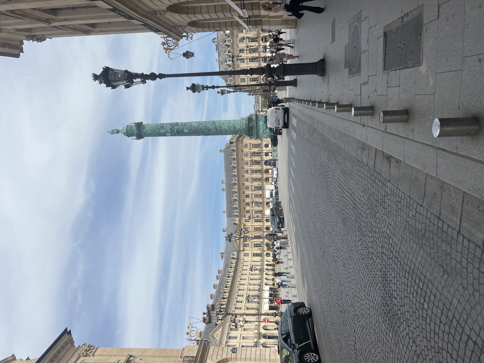 The image captures a vibrant street scene in Paris, centered around the iconic Vendôme Column, which stands tall with its distinctive green patina and a statue at its summit. The perspective appears to be from a building overlooking the street, looking down its length.
On either side of the wide street, ornate, classical-style buildings with multiple stories, numerous windows, and decorative cornices stretch into the distance, characteristic of Parisian architecture. Several vintage-style black lampposts with multiple lights line the thoroughfare, adding to the historic ambiance.
The street itself is paved with cobblestones, while the sidewalks are paved with large, light-colored flagstones. A row of silver metal bollards separates the sidewalk from the road on the right side of the image. Several people are visible on the sidewalks, walking or standing, suggesting a typical day in the city. Vehicles, including a white van and a black car (which appears to be a G7 taxi due to a visible green "G7" logo), are present on the road, indicating traffic and parked cars. Manhole covers are also visible on the sidewalk.
The scene unfolds under a bright, light blue sky with wispy clouds, suggesting a clear day, likely late morning or early afternoon. The overall impression is that of a bustling yet elegant Parisian square or avenue. No other discernible text is visible besides the G7 logo and a small red and white sign which is unclear.