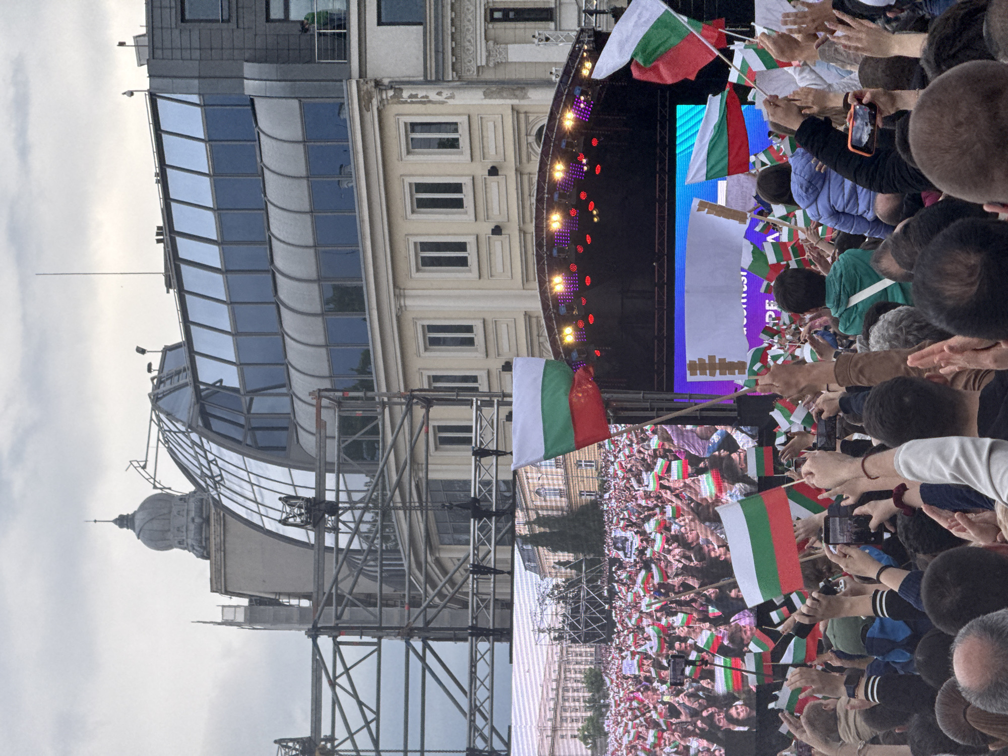 A crowd of people wave Bulgarian flags in front of a stage set up on a city square, with historic and modern buildings in the background. The event appears to be taking place in Sofia, Bulgaria, considering the flags and architecture. The scene depicts a lively gathering with visible stage lighting and scaffolding, suggesting a concert or public celebration. Many individuals are holding their phones up, likely recording the event. The buildings in the background showcase a mix of architectural styles, with a modern glass structure and older, more traditional designs. The sky is overcast, indicating possible unfavorable weather conditions.