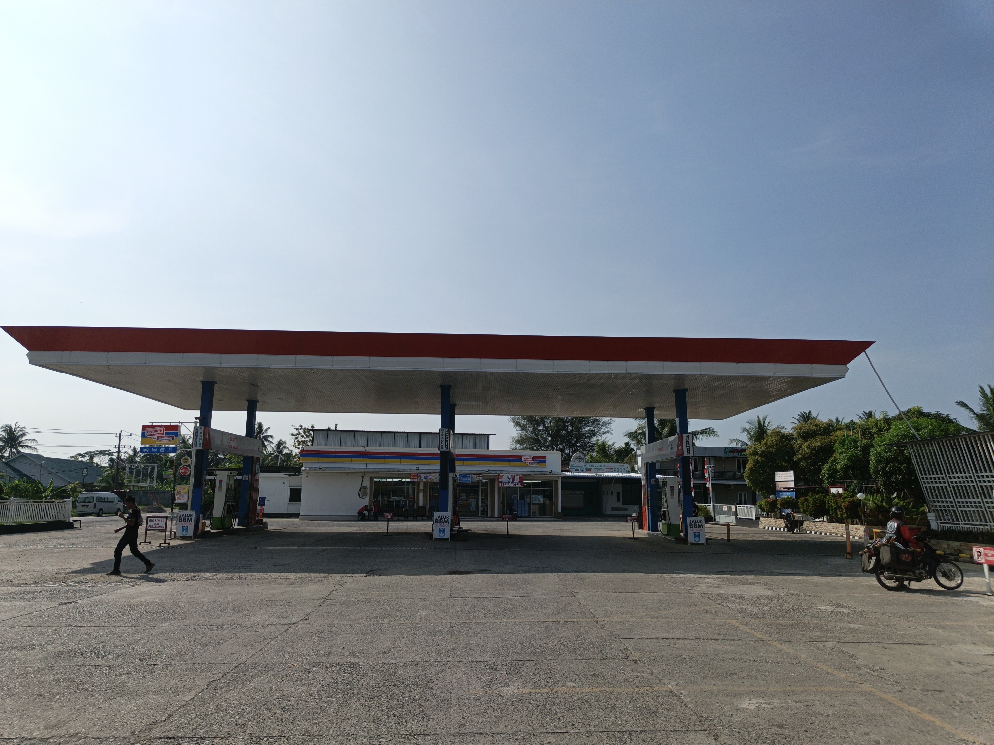 A wide-angle shot shows a gas station in Keudeteupinpunti, Indonesia, under a clear, bright sky. The prominent red-roofed gas station canopy dominates the frame. A person is walking towards the left side of the image. A motorcycle with a rider is on the right. In the background are buildings with shops. The foreground is a broad concrete surface.