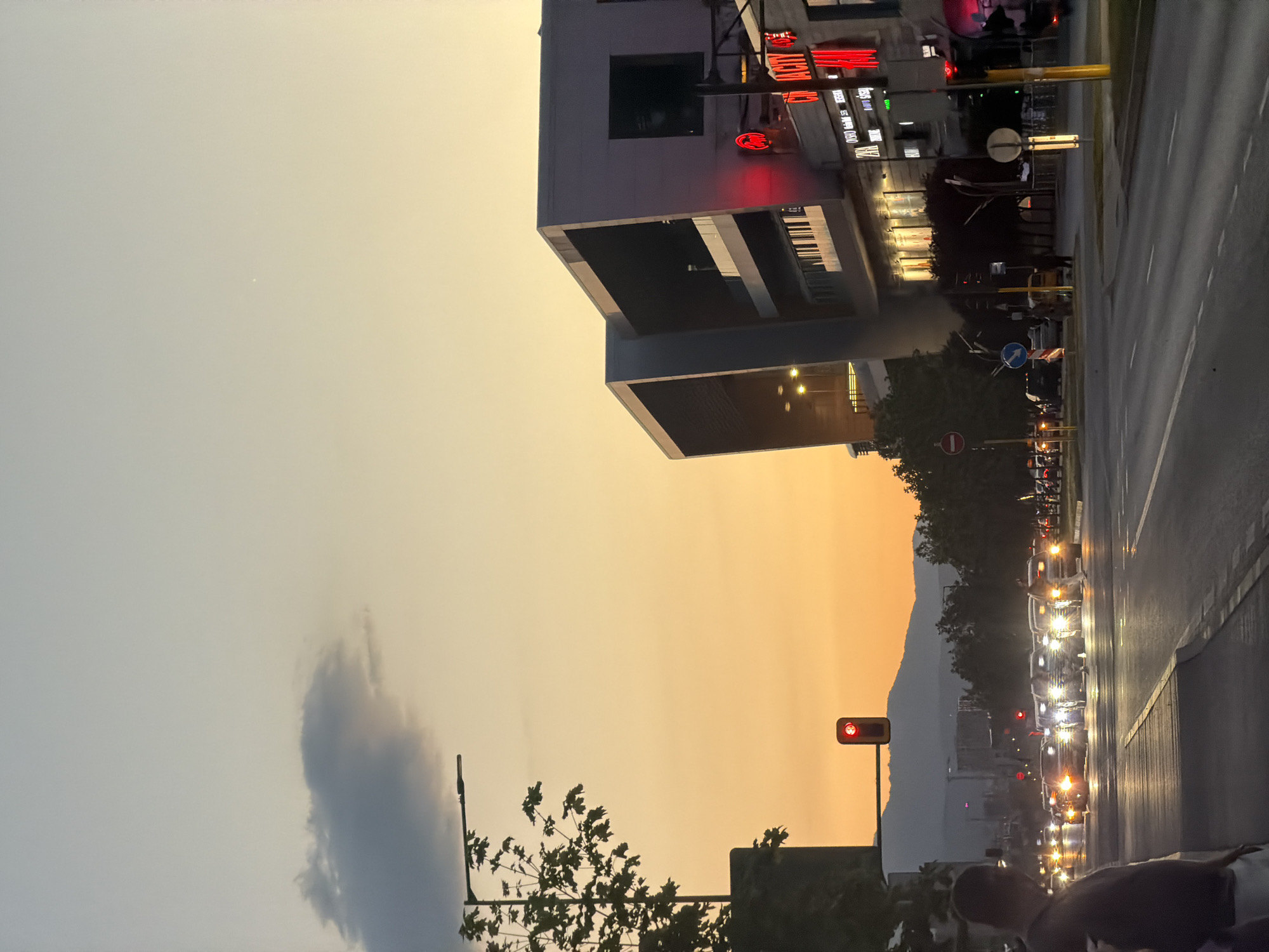 The image depicts an urban scene at dusk in Sofia, Bulgaria. The sky transitions from a light gray-blue to an orange hue at the horizon, suggesting either sunrise or sunset. The most prominent structure is a modern building with cantilevered elements and a piano keyboard graphic on its facade. Street signs and traffic lights indicate a busy thoroughfare, with numerous cars visible, their headlights blurring into streaks of light.
A mountain range is visible in the distance. Trees line the road, adding a natural element to the urban environment. A solitary cloud is seen on the left side of the image. The scene seems to be captured on a busy street in Sofia, as suggested by the architecture, traffic, and geographical context.
