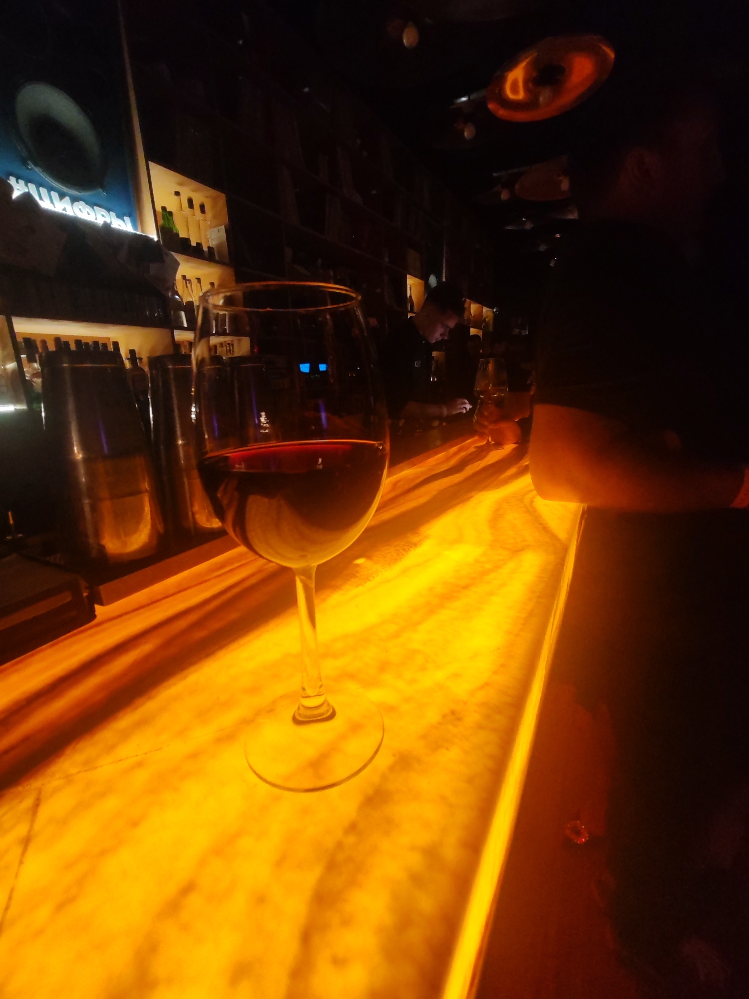 A close-up, low-angle shot captures a glass of red wine on a brightly lit, orange-hued bar surface. The glass is the primary subject, with the wine's deep red color contrasting against the warm illumination of the bar. In the background, the scene is dimly lit and suggests a bar or restaurant setting. Several metal shakers are visible behind the wine glass, lined up on a shelf. Further back, a wall adorned with shelves holds numerous bottles, indicating a well-stocked bar. Above the bottles, a large, circular object is mounted on the wall, with white text in an Cyrillic script underneath it. In the far background, a person is partially visible, appearing to be a bartender or server, looking down and possibly interacting with a phone or tablet. The overall mood is atmospheric and intimate, with the warm lighting creating a sense of ambiance typical of a nighttime establishment. The focus is on the wine and the inviting glow of the bar, hinting at a moment of relaxation or enjoyment.