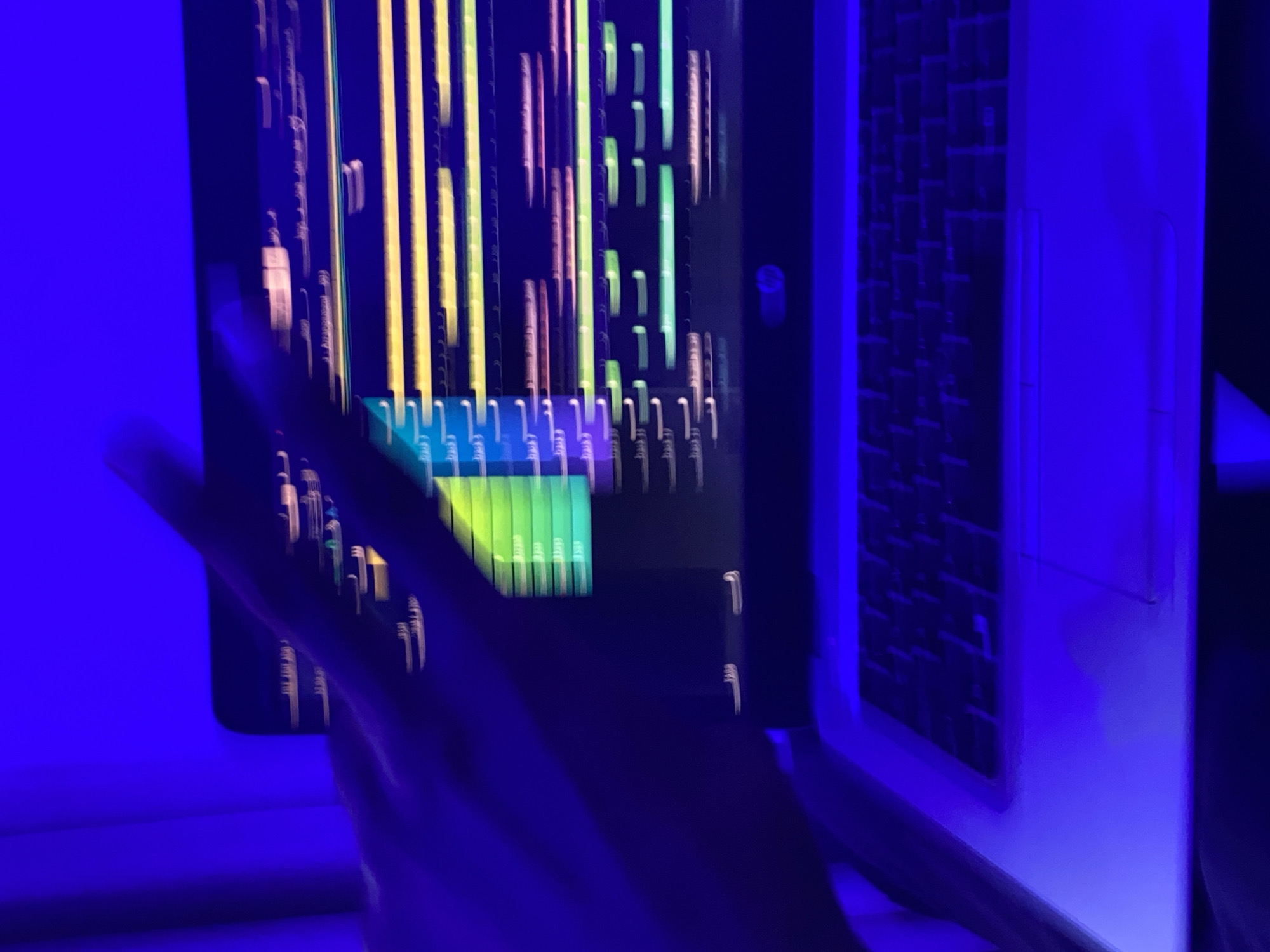 The blurry, low-light image captures a workstation bathed in intense blue illumination, likely in Ibadan, Nigeria. The central subject is a laptop screen displaying a dynamic, vertically arranged graphic composed of numerous colorful bars in shades of yellow, green, pink, and blue. This display strongly resembles an audio waveform, a timeline, or data visualization often seen in a Digital Audio Workstation (DAW).
A blurred human hand, illuminated by the pervasive blue light, reaches into the frame from the lower left, appearing to interact with or gesture towards the laptop screen. To the right, the open keyboard and chassis of another laptop are partially visible, featuring dark keys against a lighter background, all bathed in the same blue glow.
The strong, artificial blue lighting suggests the scene takes place at night or in a deliberately darkened room with specialized mood lighting. While much of the detail is obscured by blur, faint, small white text is visible beneath some of the colored bars on the screen. This text appears repetitive and could be time indicators like "00:00" or possibly Arabic script, such as "الوقت" (meaning 'the time'), which is indistinct due to the image quality. No other specific location details or emotional cues are discernible from the image.