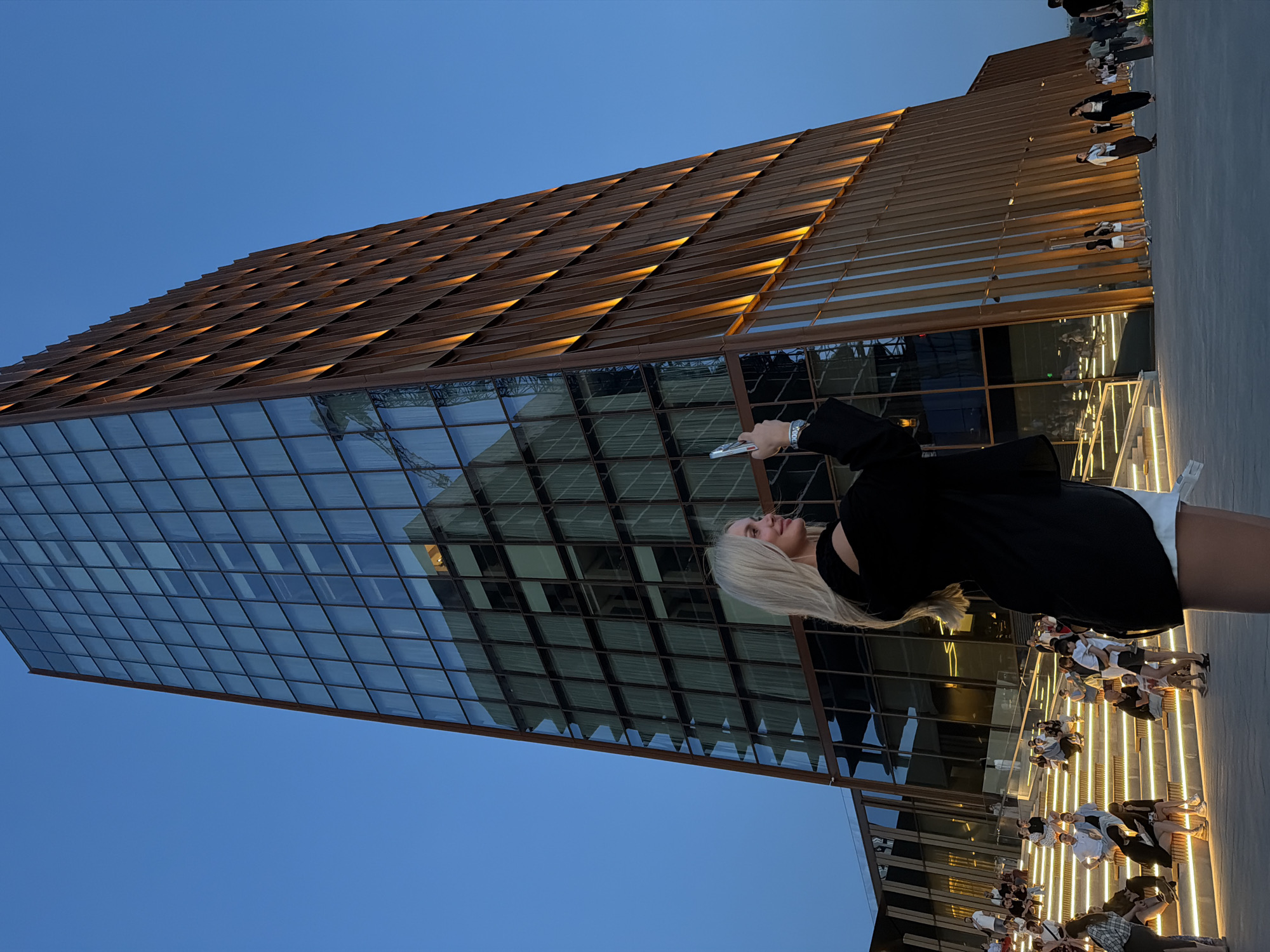The image depicts a woman with blonde hair standing in front of a modern building in Kazan, Russia. The building features a glass facade with a grid-like pattern, topped by a structure with horizontal wooden elements illuminated from within. People are seated on benches along the base of the building. The sky is a clear blue. The woman is holding a phone.