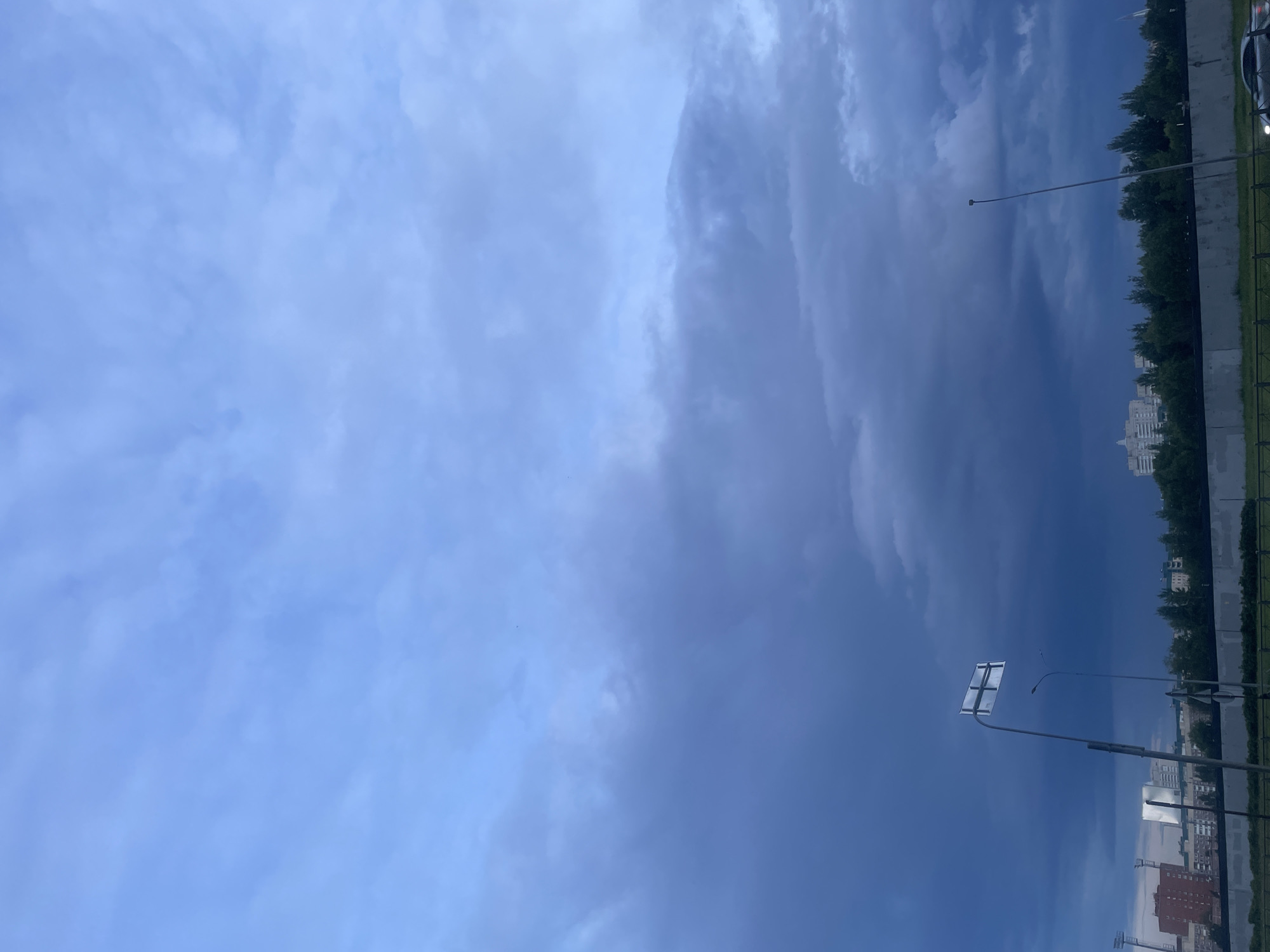 A low-angle, full shot captures a dramatic, overcast sky above a city skyline in Saint Petersburg, Russia. The sky is a deep, bruised blue with heavy, dark clouds dominating the lower half of the frame, suggesting an approaching storm. Above, lighter, wispy clouds are illuminated by diffused sunlight. In the mid-ground, a line of trees separates the sky from the urban landscape. Several buildings, varying in height and architectural style, are visible behind the trees, with one tall, modern building standing out. A dark, concrete barrier runs horizontally across the foreground. Two streetlights and a traffic sign are prominent on the left side of the image. A silver car is partially visible on the right, its headlights on, suggesting movement. The overall mood is somber and foreboding due to the dark clouds.