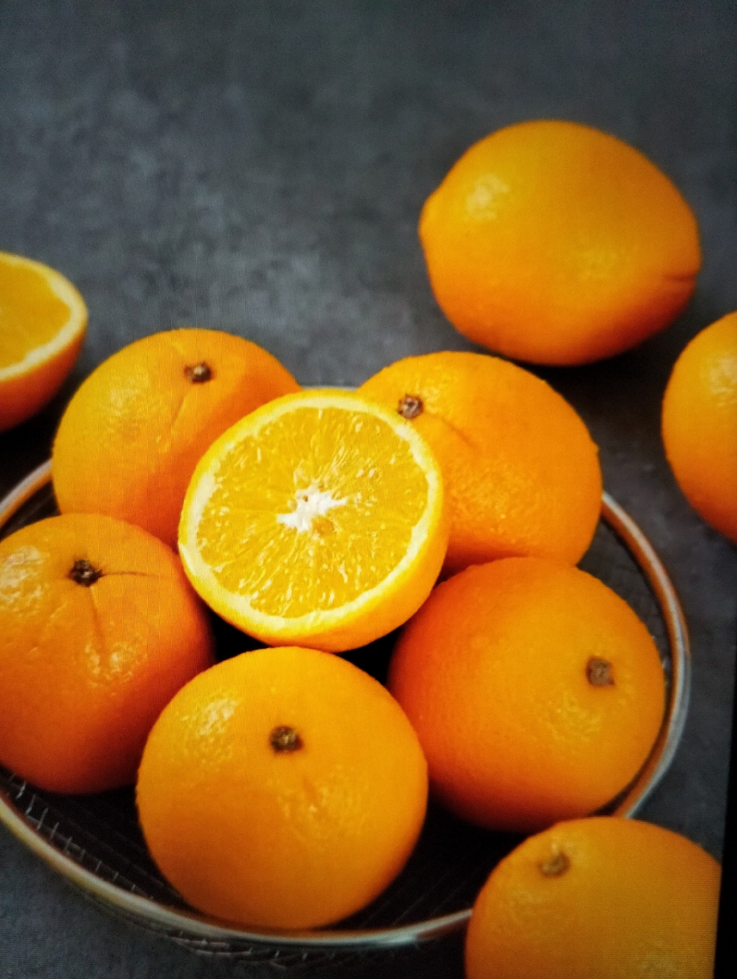 A shallow depth of field shot shows a close-up of several whole and sliced oranges arranged on a dark gray surface. The oranges are a vibrant orange color, with some showing small brown stem marks. One orange is cut in half, revealing the segmented interior and white pith. The oranges are partially contained within a round, silver wire basket. The background is a dark, textured surface, possibly a stone or concrete countertop. The lighting is soft and diffused, highlighting the textures and colors of the fruit. There are no people or specific landmarks visible in the image, nor is there any indication of a specific location within Payagaboh, Indonesia.