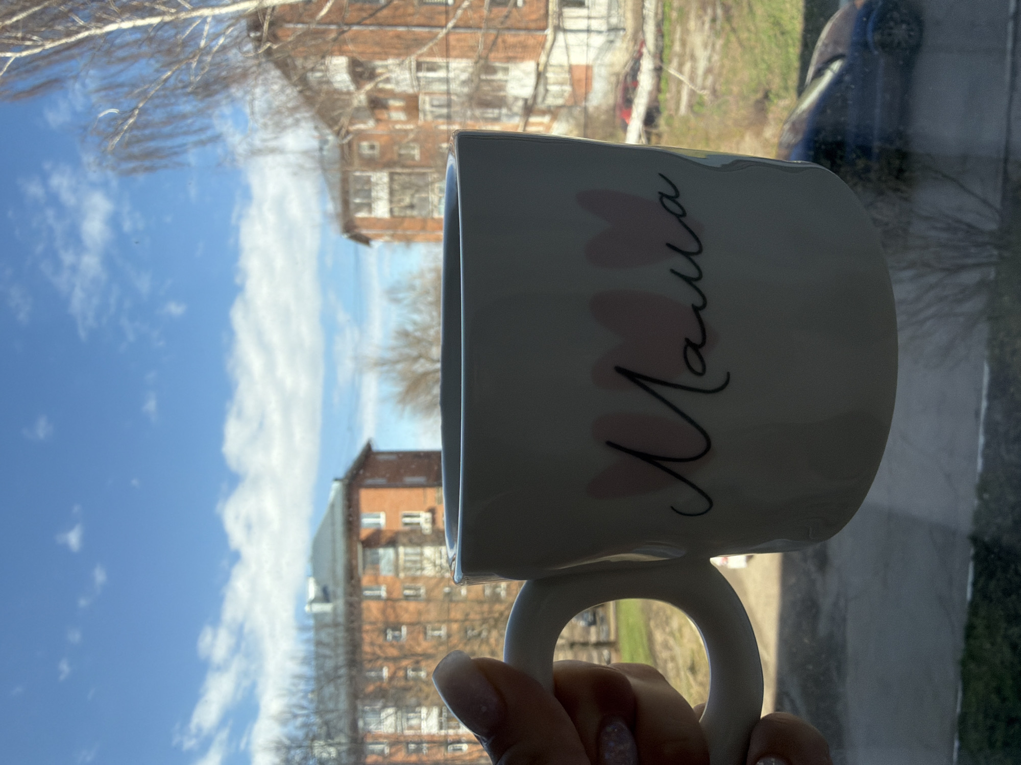 A mug with the word "Mama" written in Russian cursive is held up in front of a window. The window shows a view of multi-story brick buildings under a blue sky with white clouds. The location is Votkinsk, Russia. A hand with painted fingernails holds the mug. Reflections of a car and surrounding green space are visible in the window glass.
