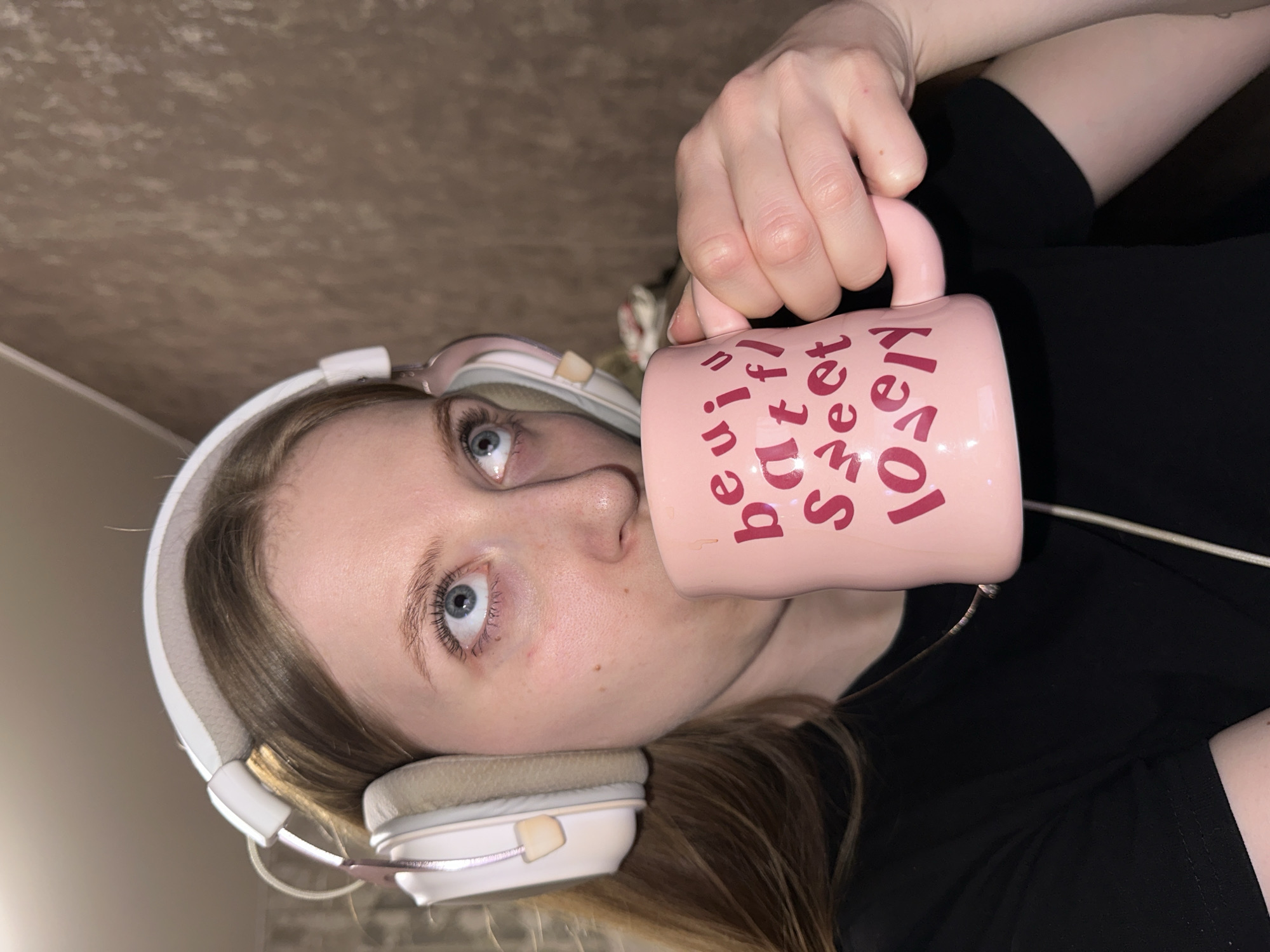 The scene is depicted indoors in the city of Chaikovsky, Russia. A female individual with light brown hair, wearing a black short-sleeved t-shirt and white over-ear headphones with beige earcups, is present. A thin white audio cable descends from the headphones. The individual holds a pink ceramic mug, inscribed with red text partially visible as "beautiful sweet lovely," to her lower lip, indicating consumption of a beverage. Her blue eyes are directed upwards and slightly to the right. The background consists of a light brown wall featuring a textured pattern.