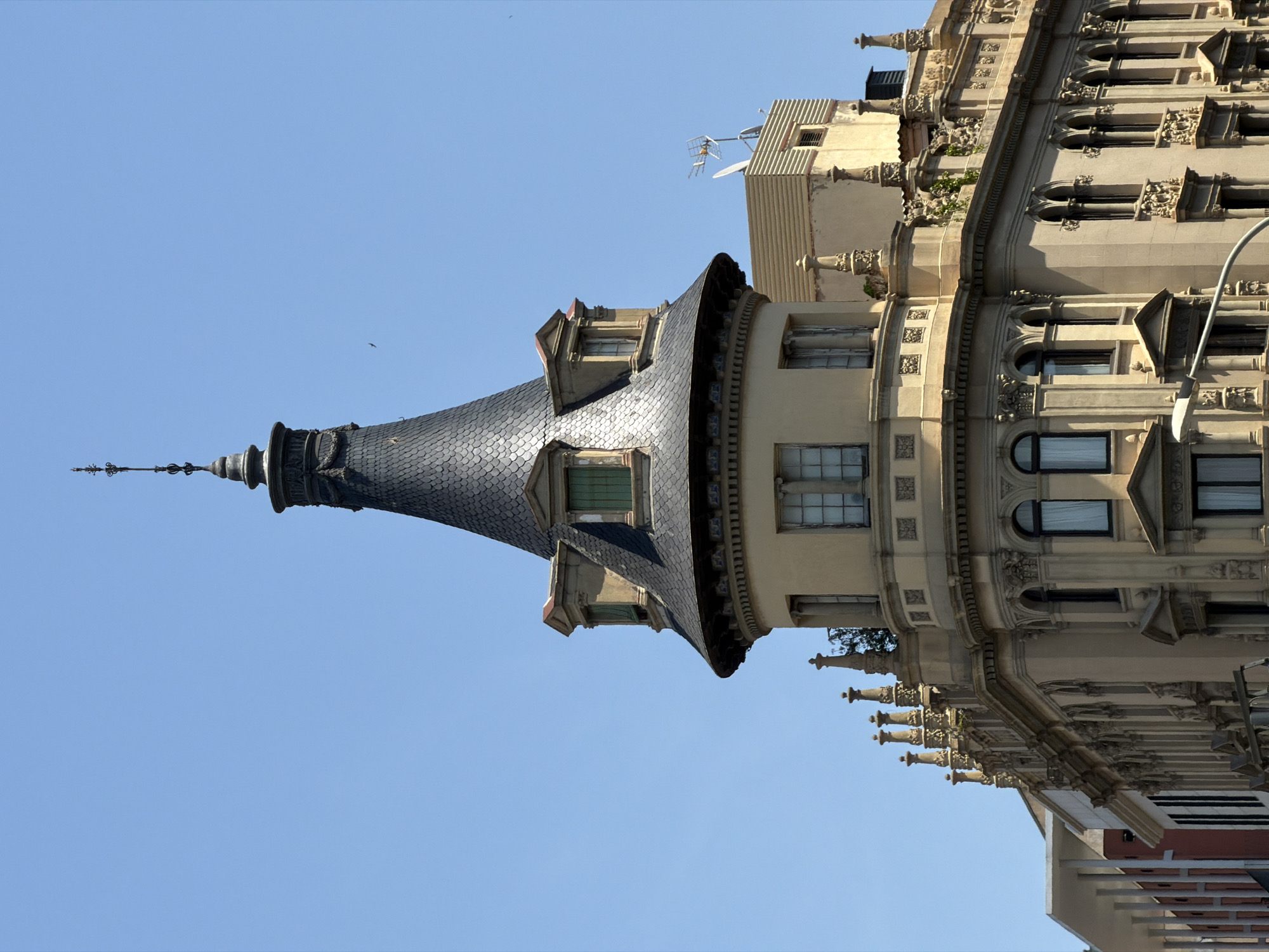 This image shows a close-up, low-angle view of the upper facade of a multi-story building in Barcelona, Spain, under a clear blue sky. The building exhibits ornate architectural details, including a prominent conical tower with dark gray, shingle-like roofing and decorative finials. The tower features dormer windows with green shutters and sits atop a curved section of the building. Below the tower, the facade is adorned with classical architectural elements such as pilasters, decorative moldings, and arched windows. The main body of the building is a light beige color, contrasting with the darker roof of the tower. Other buildings are visible in the background, including one with a ribbed roof and television antennas. The image appears to have been taken on a sunny day, casting distinct shadows on the building. There are no people or vehicles visible in the frame.