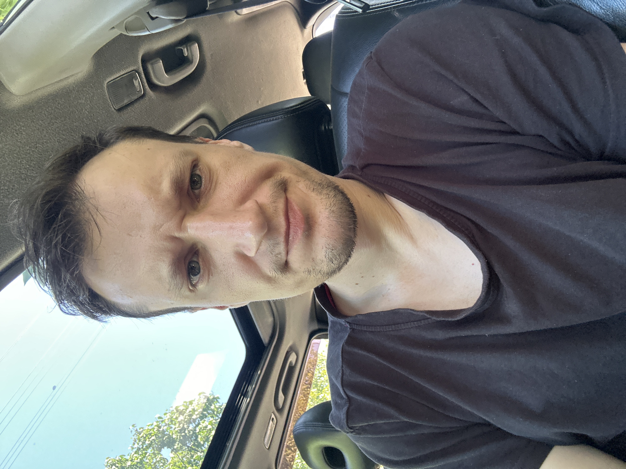 A male with light skin and brown hair is sitting in the driver's seat of a car, looking at the camera. He is wearing a dark t-shirt. His eyebrows are furrowed, and he has a slight stubble. The car has a sunroof, which is open, revealing a bright blue sky with some trees visible in the background. The interior of the car is visible, including a black headrest. The image appears to be a selfie taken from inside the vehicle. There is no information available that indicates this image is located in Makiv, Ukraine.