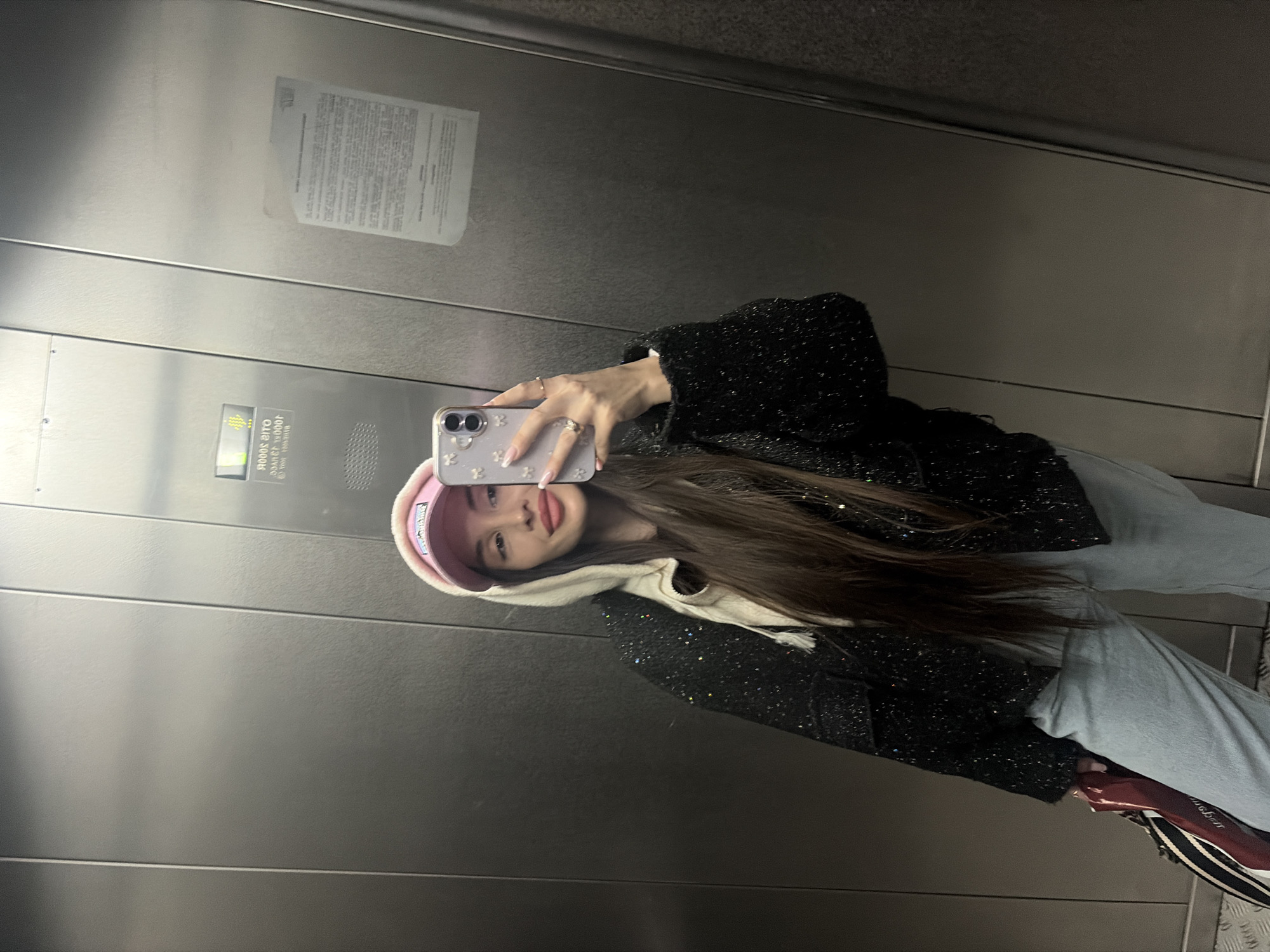 A young woman with long brown hair is taking a selfie in a metal elevator. She is wearing a pink visor cap, a hooded jacket, and light-colored jeans. Her phone has a decorative case. The elevator walls are metal with horizontal seams and some signage. The photo was taken indoors. There is no indication the photo was taken in Saint Petersburg, Russia.