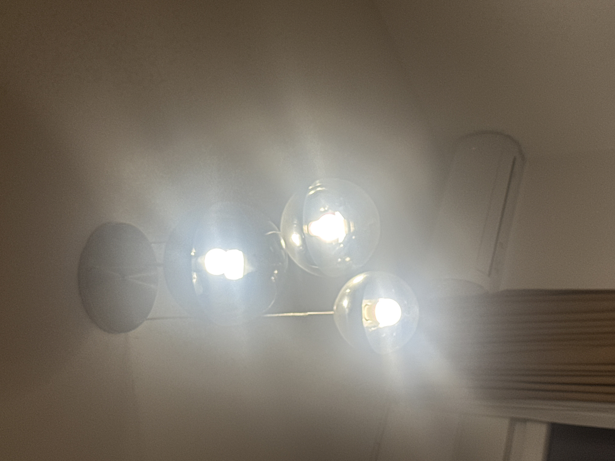 The image shows a close-up of a modern chandelier with multiple glass globe lights. The lights are on and emit a bright, white glow, creating a diffused lighting effect that obscures some of the details. The chandelier is suspended from a ceiling or wall, and the background is a plain, neutral color, likely a wall or ceiling. To the right of the chandelier, a portion of what appears to be window blinds is visible, suggesting the setting is indoors, possibly in a home or office. There are no people visible in the image. The overall impression is one of contemporary design and ambient lighting. The lack of specific details about the surroundings makes it difficult to pinpoint the exact location beyond it being an interior space. The time of day and weather are not discernible. There is no visible text in the image.
