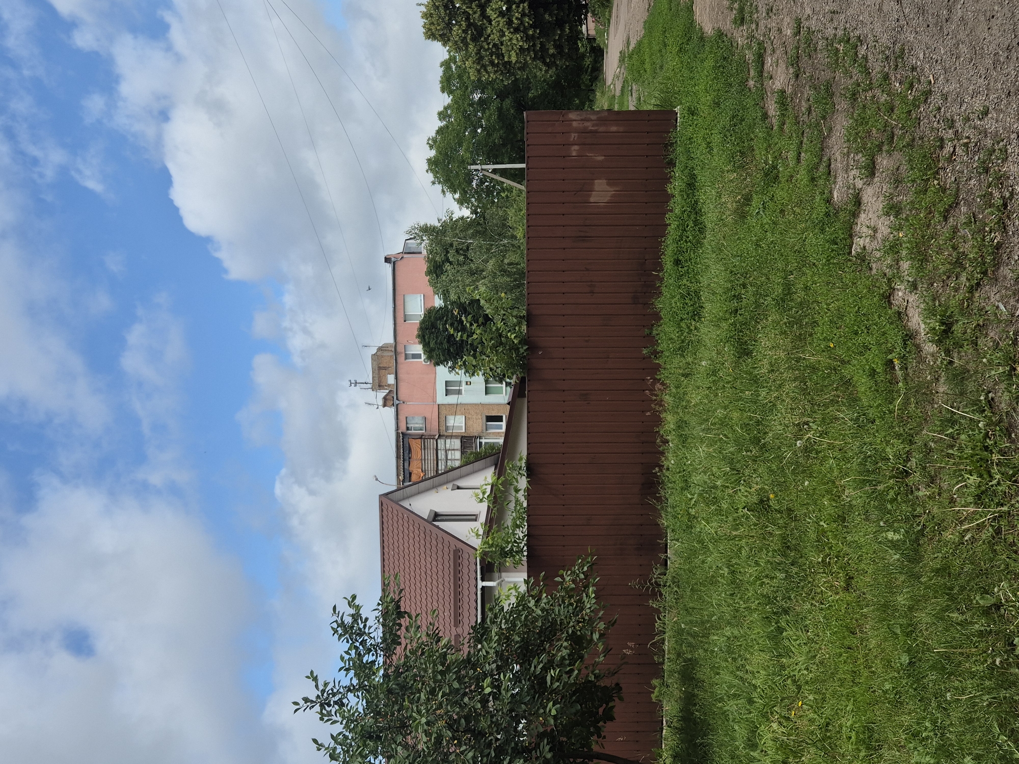 This is a daytime, medium-angle, outdoor shot of a rural scene in Sozonivka, Ukraine. A tall, reddish-brown corrugated metal fence dominates the foreground, stretching horizontally across the frame. To the left of the fence, a lush green tree with dark leaves is partially visible. Beyond the fence, a white house with a reddish-brown tiled roof is situated. In the background, a multi-story building, painted in pink and light blue, stands amidst a cluster of green trees. The sky above is blue with scattered white clouds. A dirt path runs along the right edge of the image, bordering the fence and extending into the distance. The ground in front of the fence is covered in green grass.