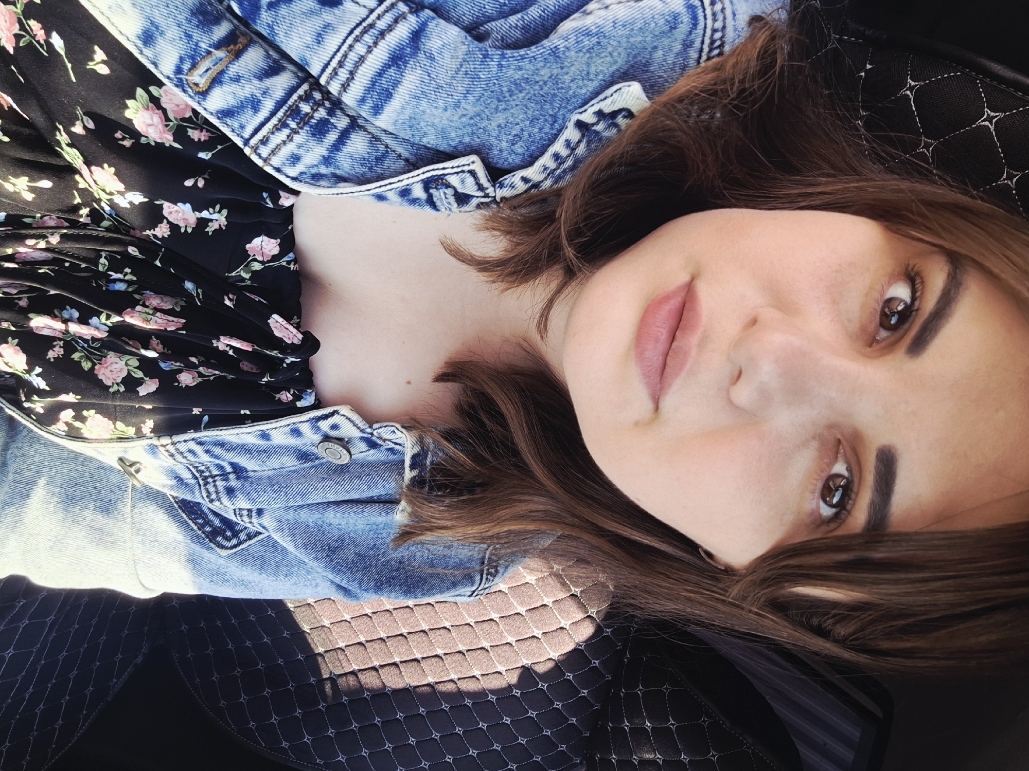 A woman is lying on a surface, possibly a car seat, wearing a floral dress and a denim jacket. She is facing the camera. The location is unknown and cannot be determined from the image.