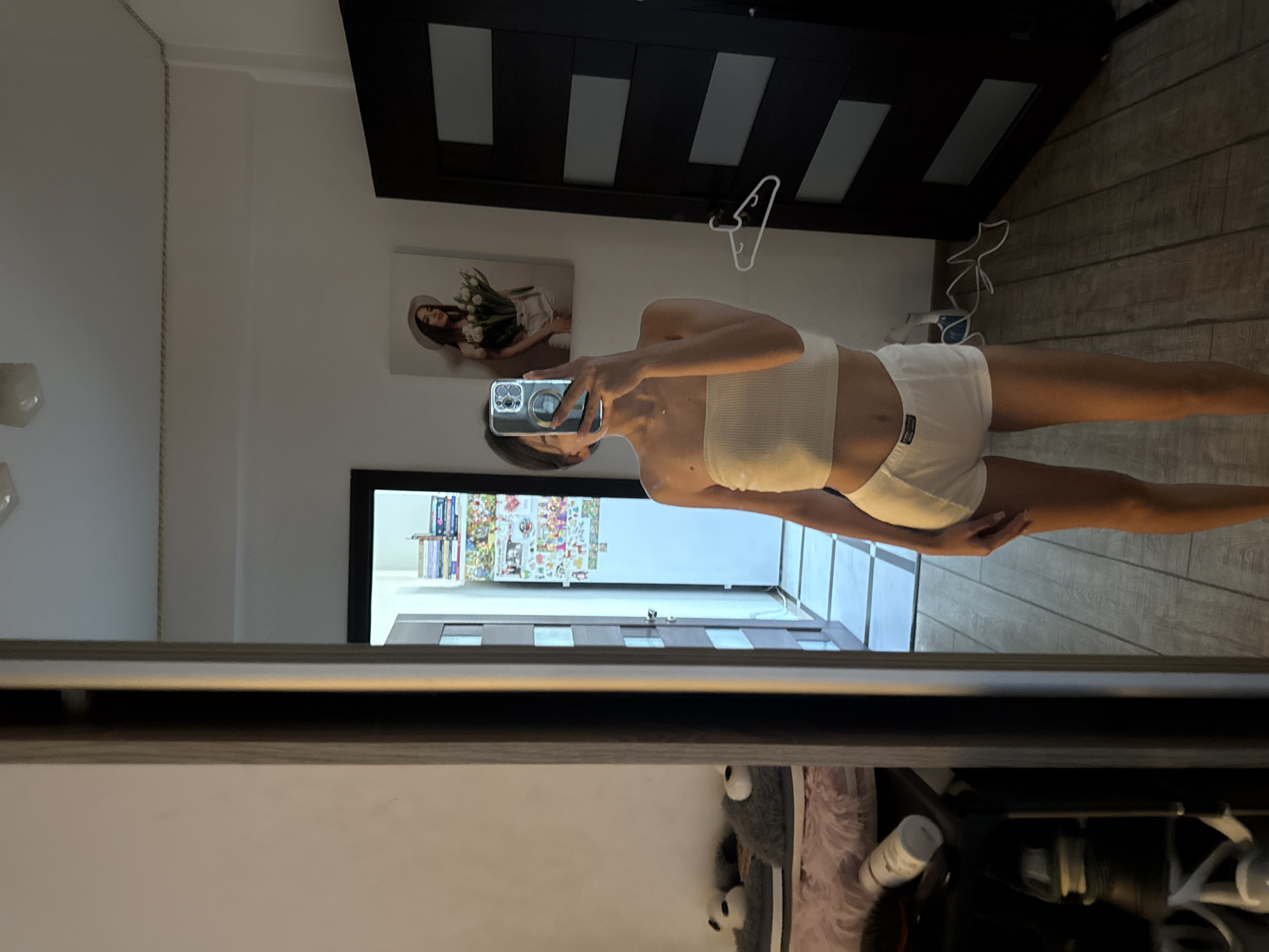 The image depicts a young woman taking a mirror selfie in what appears to be a bedroom or dressing area. She is wearing a white strapless top and white shorts. The room has light-colored walls and wooden flooring. A large mirror is central to the image, reflecting the woman and a portion of the room behind her. To the left of the mirror, a doorway leads to another area with a closed door and what looks like a shelf with various items. In the reflection, a painting of a woman holding flowers hangs on the wall. A closed door with a patterned design is visible on the right side of the image. The scene suggests a personal, indoor setting. There is no visual information to ascertain the specific location within Chișinău, Moldova.