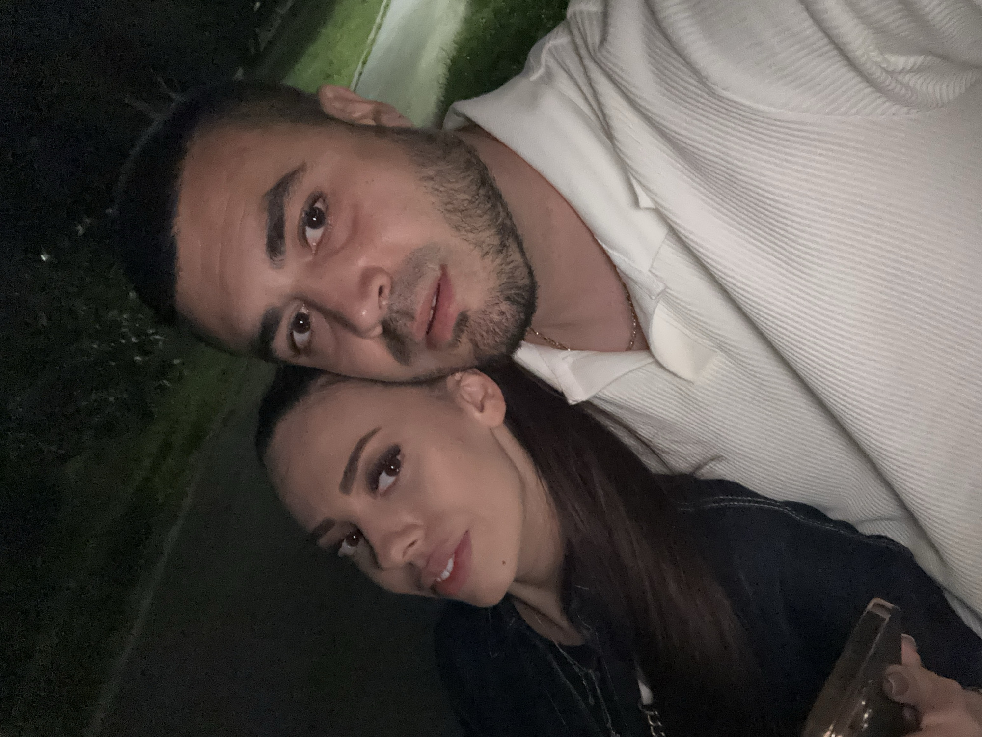 The image displays a head-and-shoulders selfie of two individuals, an adult female and an adult male, captured in a dimly lit environment, likely outdoors at night, with illumination focused on their faces.
The female is positioned on the left, with her head slightly tilted to her right, resting near the male's shoulder. She has dark, long, straight hair and is wearing dark clothing, possibly a jacket. Her right hand is visible in the lower left corner, holding a small, dark, rectangular object, possibly a smartphone. She is looking towards the camera with a slight smile.
The male is on the right, directly facing the camera with a neutral expression. He has short, dark hair with faded sides and a short beard/stubble. He is wearing a light-colored, possibly white or off-white, collared shirt with a distinct vertical ribbed texture. A thin, gold-colored chain necklace is visible around his neck.
The background is dark and largely out of focus, showing indistinct dark shapes consistent with trees or bushes and faint patches of green, suggestive of grass or foliage. A lighter, blurry area in the upper right background might indicate a pathway or distant illuminated surface.
The image contains no discernible features or landmarks to confirm the location as the city of Varna, Bulgaria.