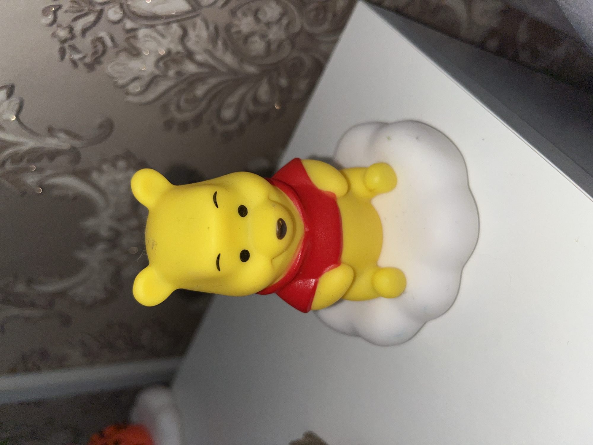 A yellow Winnie the Pooh toy figure, wearing a red shirt, sits on a white cloud-shaped base. The figure is positioned in the center of the image and is made of plastic. The background consists of patterned wallpaper and a white surface, likely a table or shelf. The lighting suggests an indoor environment. There is no information in the image to suggest a location in Sharan, Russia.