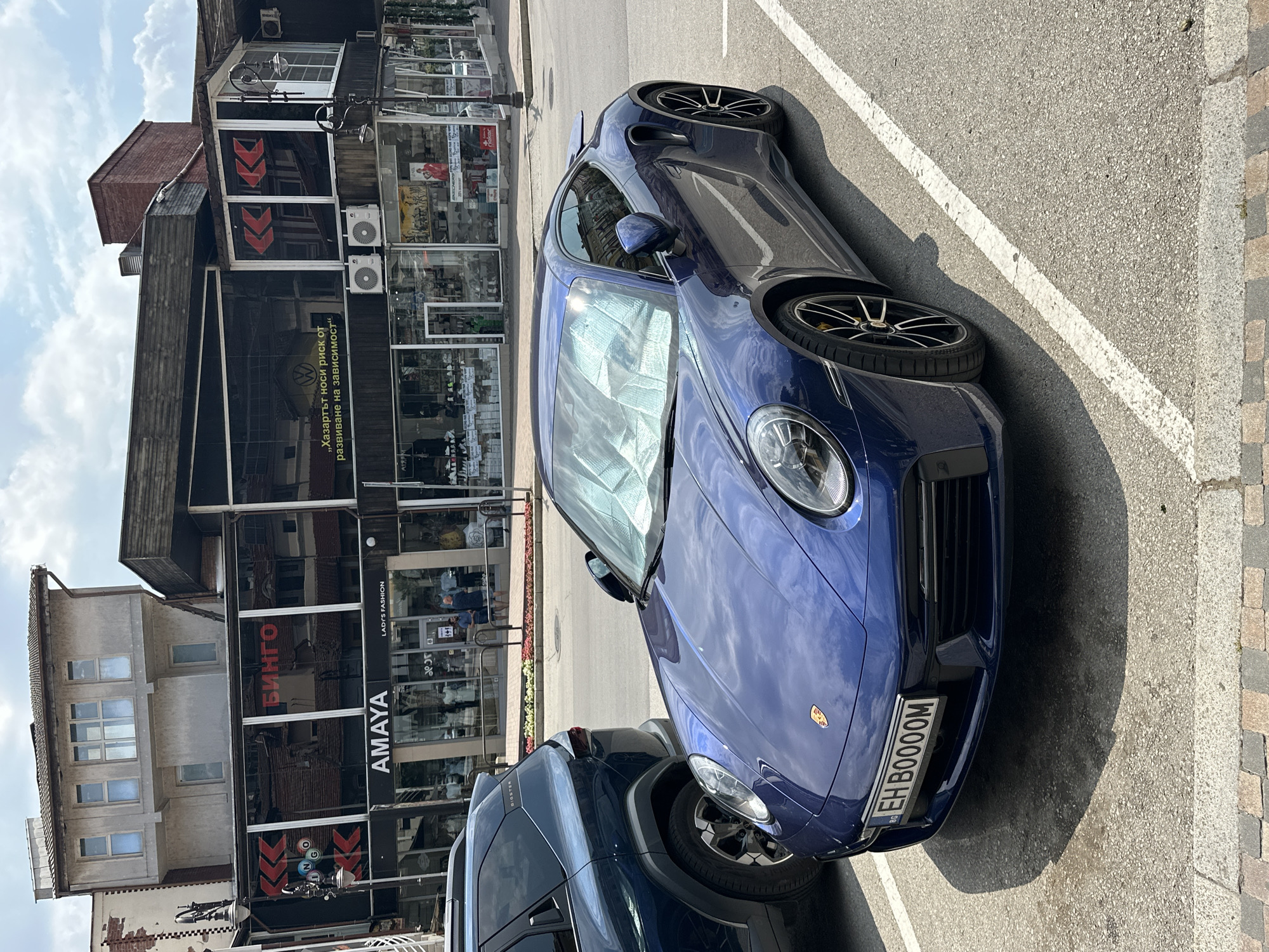 This vertical, eye-level shot captures a blue Porsche 911 parked on an asphalt surface with a white line marking a parking space. The car is the primary object, angled slightly towards the camera, revealing its sleek design and distinctive headlights. The license plate, "EH B0000M", is clearly visible on the front.
Behind the car, a multi-story building with a mix of modern and older architectural elements stands. The ground floor of the building features storefronts with large glass windows, displaying various merchandise and signage. Notable text visible on the storefronts includes "AMAYA LADY'S FASHION" and "БИНГО". There is also a sign with the text "„Хазартът носи риск от развиване на зависимост" which translates to "Gambling carries a risk of developing addiction". The building's facade shows signs of wear and tear on the upper levels, contrasting with the more modern retail spaces below.
The scene appears to be a street in Veliko Tarnovo, Bulgaria, given the Cyrillic text on the signs and the overall urban environment. The lighting suggests it's daytime, with bright sunlight casting shadows, indicating fair weather. No people are clearly visible in the immediate vicinity of the car, though blurry figures can be seen through the shop windows. The overall impression is a quiet street scene with a focus on the luxury car against a backdrop of commercial establishments.