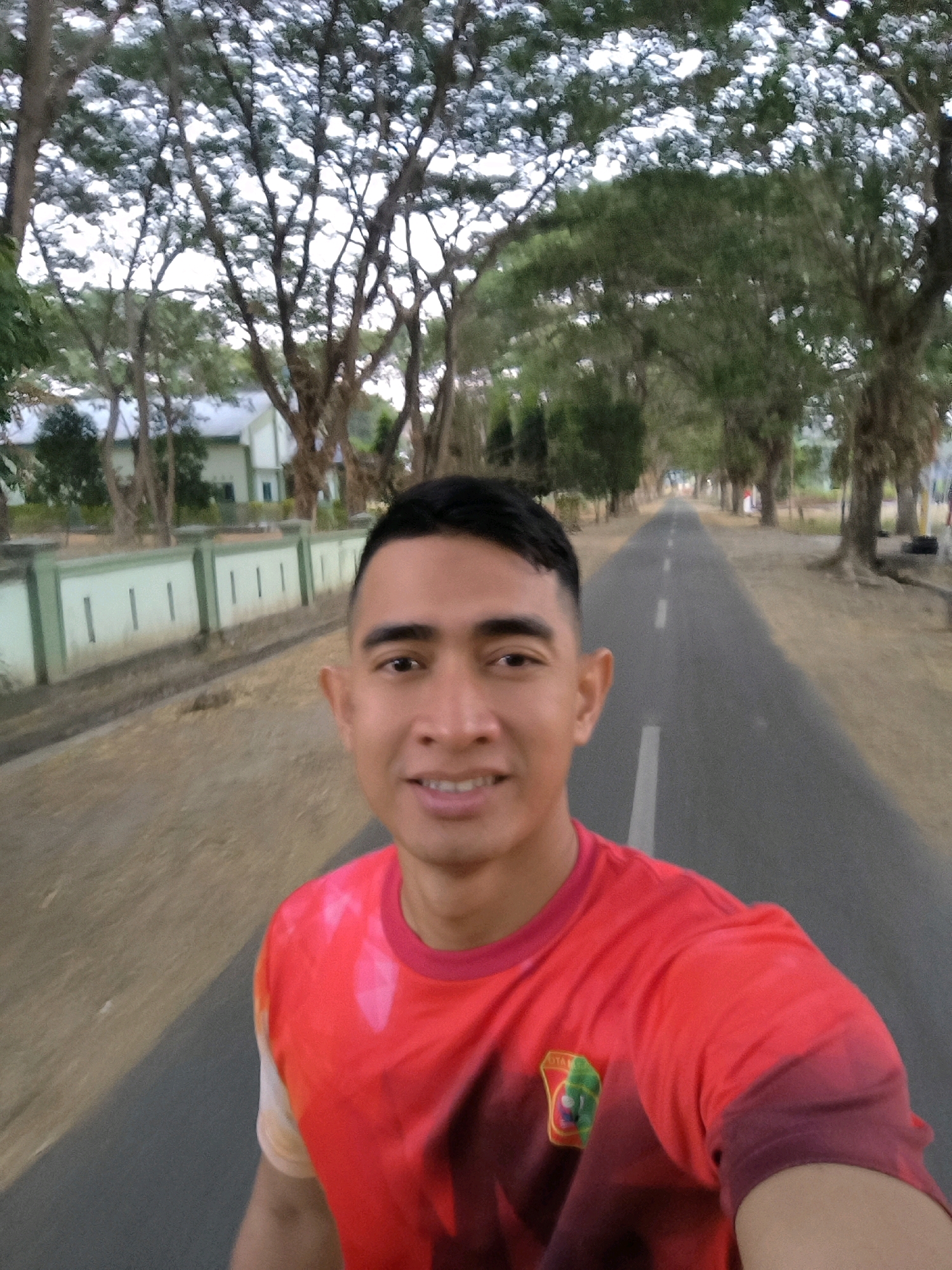 A man with short dark hair and a light brown complexion is shown from the chest up, smiling and looking directly at the camera. He is wearing a red t-shirt with a circular logo on the left side of his chest. The man is on a paved road, which curves to the right and disappears into the distance. Trees with full canopies line both sides of the road, creating a tunnel effect. In the background, a light green and white building with a red roof is visible behind a low, light green fence with dark green pillars. The sky is overcast and appears to be either early morning or late afternoon. The overall scene suggests an outdoor setting in a tropical or subtropical region.
