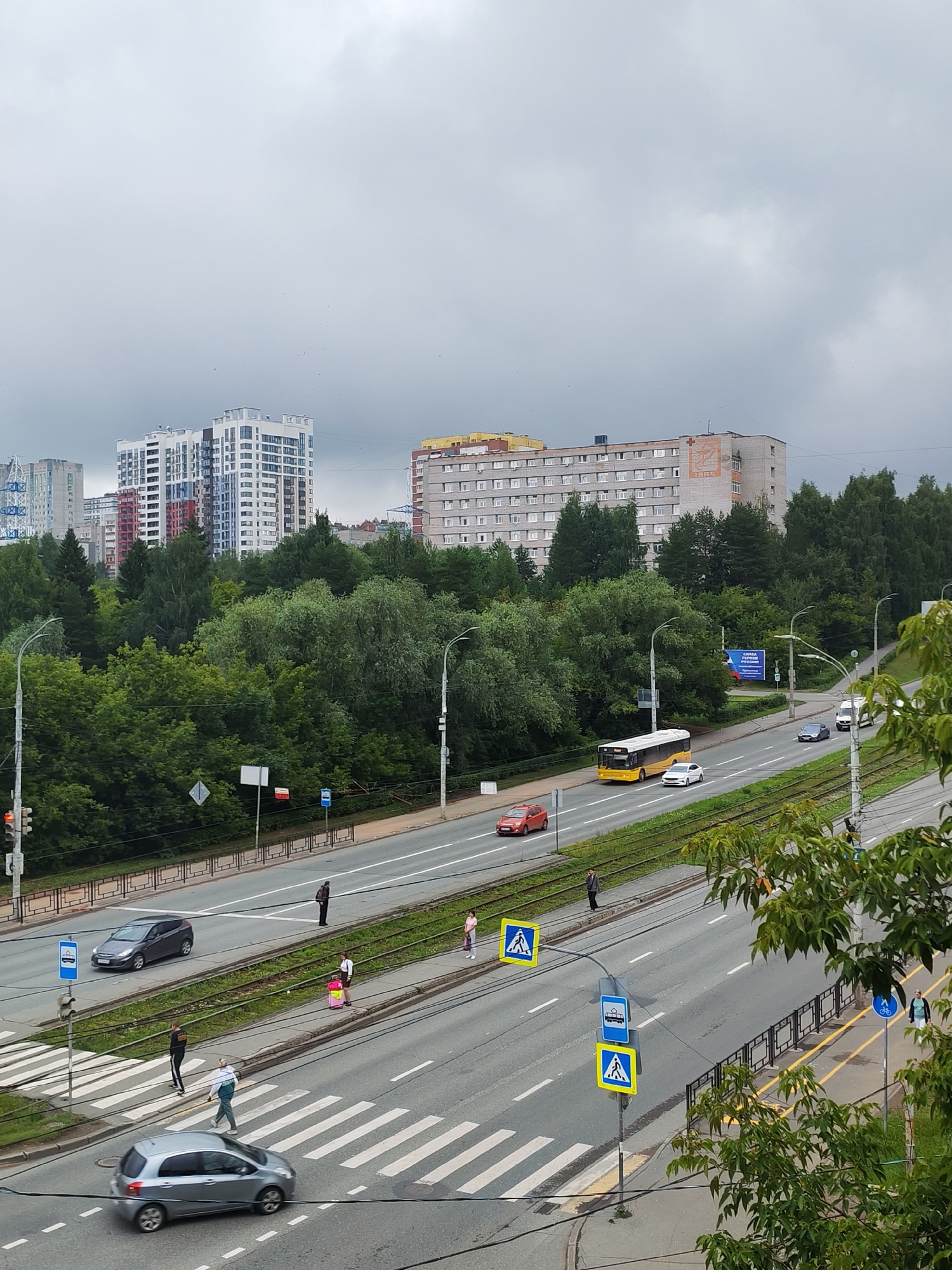 This is an aerial, eye-level, medium shot of a city street in Izhevsk, Russia, on an overcast day. The street is a multi-lane road with trolley tracks running down the median. There are several buildings visible in the background, including a tall, modern residential building and a longer, older building with a distinctive orange emblem and the year "1985" inscribed on it. A line of trees separates the road from the buildings.
On the street, there are several vehicles, including a yellow bus, a red car, a dark gray sedan, a white van, and a silver hatchback. Pedestrians are crossing the street at a marked crosswalk. One person is walking with a pink suitcase. There are also traffic lights and road signs visible. The scene appears to be a typical urban street with moderate traffic and pedestrian activity.