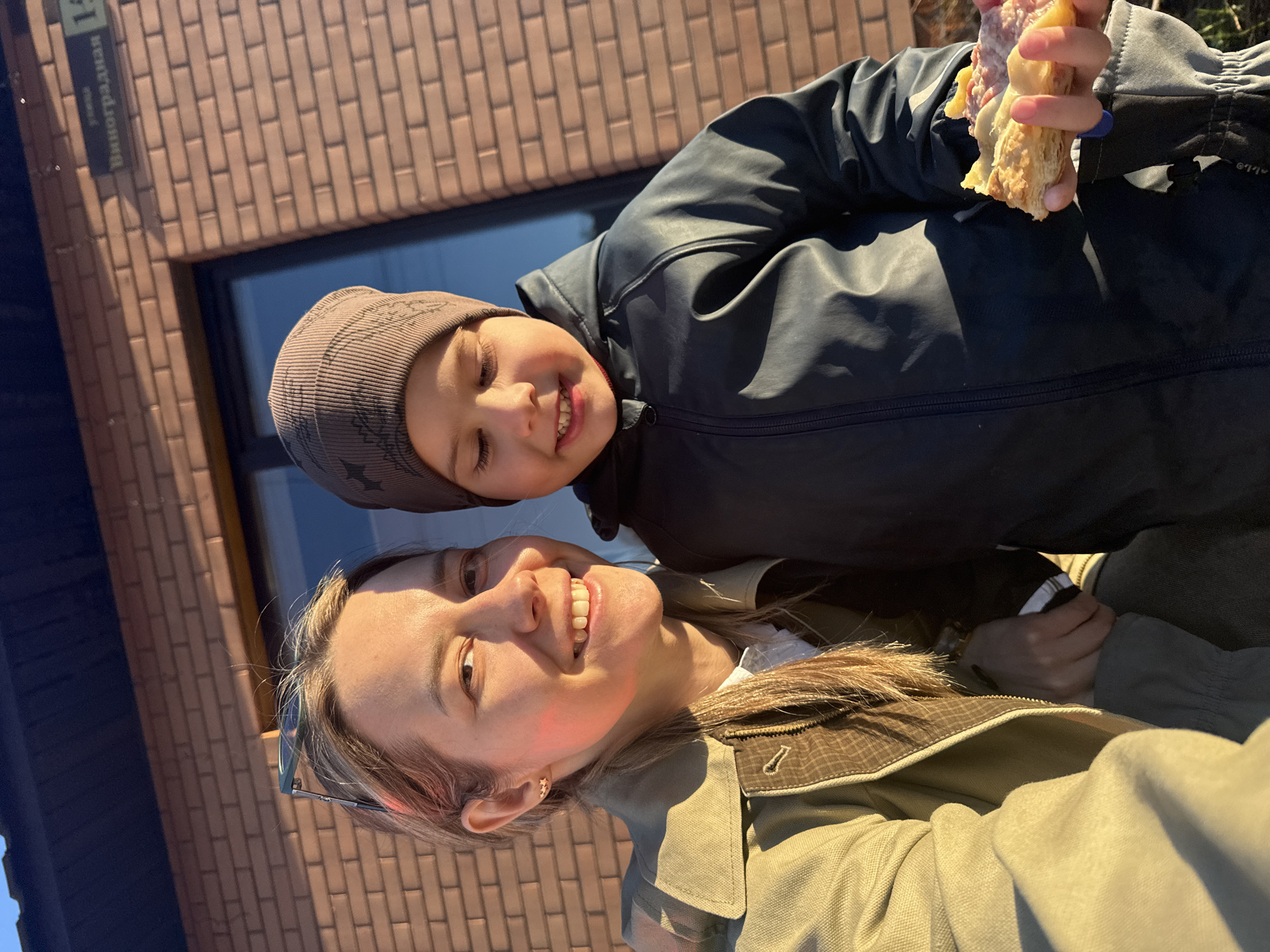 A woman and a young boy are posing for a selfie outdoors in the city of Chaikovsky, Russia, with the sign indicating the address as Vinogradnaya street, 14. The woman is smiling at the camera, wearing a light green jacket. The boy is also smiling, wearing a dark beanie with stars, a dark jacket and holding a sandwich with salami. A brick building with a window is in the background. The photo is lit by sunlight.