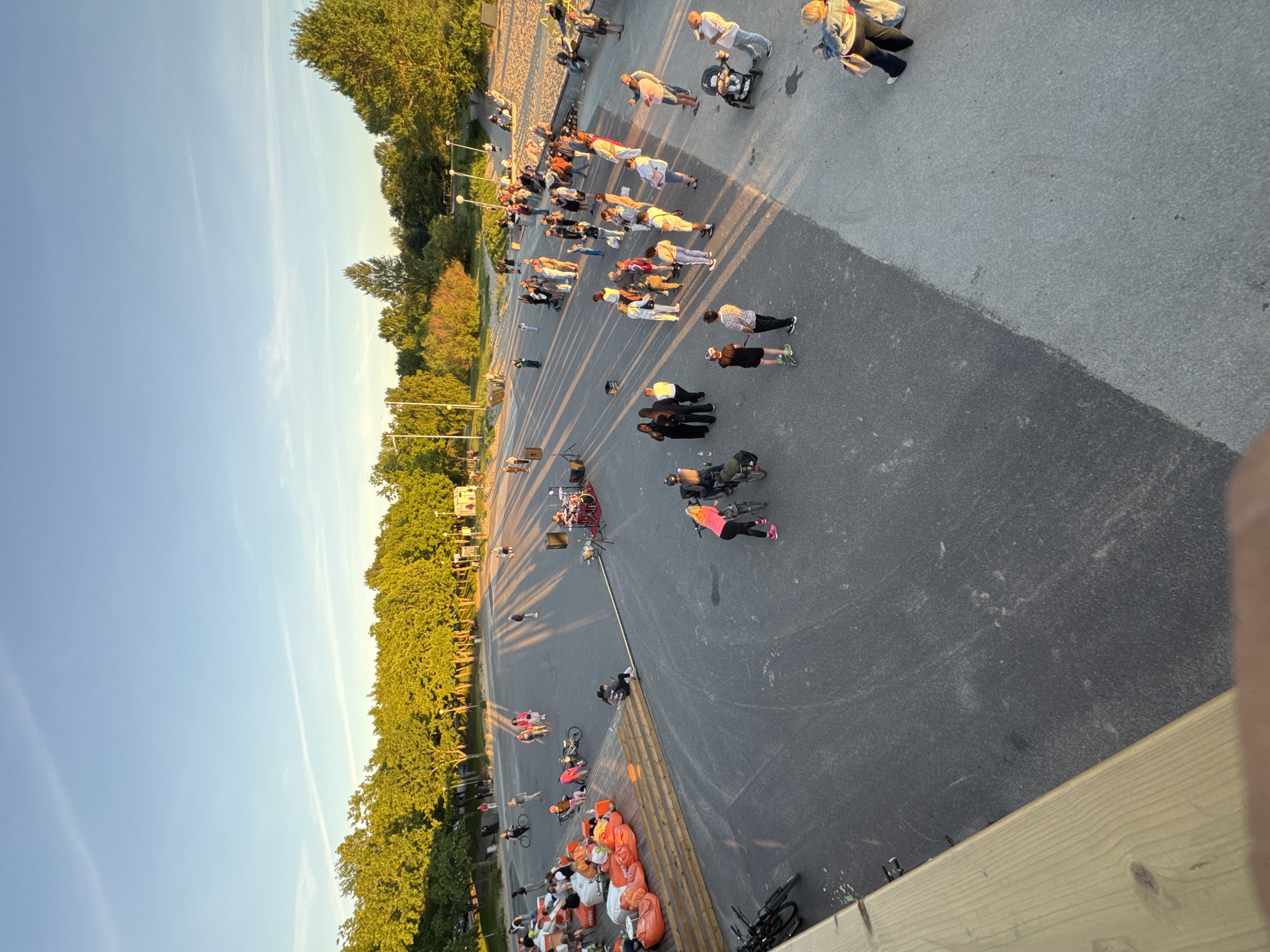 This is a high-angle, full-shot, eye-level outdoor photograph taken in Tallinn, Estonia, at sunset. The scene depicts a public gathering in an open, paved area bordered by trees and a sandy embankment. A group of people are gathered in the foreground, some seated on orange beanbag chairs on a wooden platform, while others stand or walk on the pavement. In the mid-ground, a band is set up with drums and speakers, suggesting a performance. Many individuals are engaged in conversation, observing the band, or walking with bicycles. The setting sun casts long shadows across the paved area, illuminating the trees with a warm, golden light. The sky is a clear blue with a few wispy clouds. The overall atmosphere appears relaxed and communal, with people enjoying the evening outdoors. The sandy area in the background suggests proximity to a beach or waterfront.