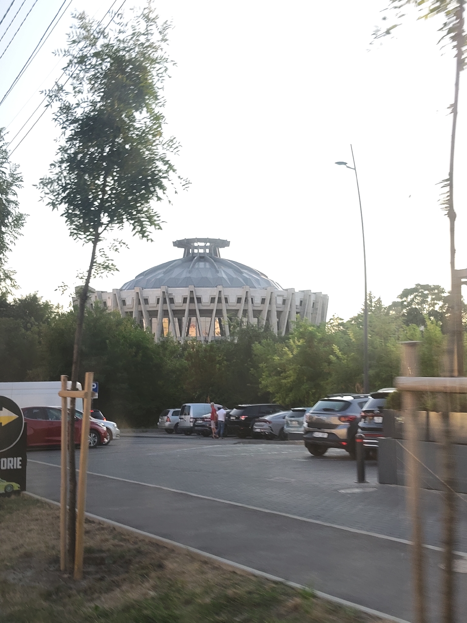 The image shows a large, circular building with a ribbed dome and a stepped roof, partially obscured by trees. The building is located in Chișinău, Moldova, and appears to be the Palace of the Republic. In the foreground, there is a parking lot with several cars, including a red hatchback, a silver SUV, and a black SUV. Two people are standing near the cars, one wearing a white shirt and the other a red shirt. A street lamp is visible in the background, and power lines stretch across the sky. The overall scene is a daytime shot, with a bright sky and dappled sunlight filtering through the trees.