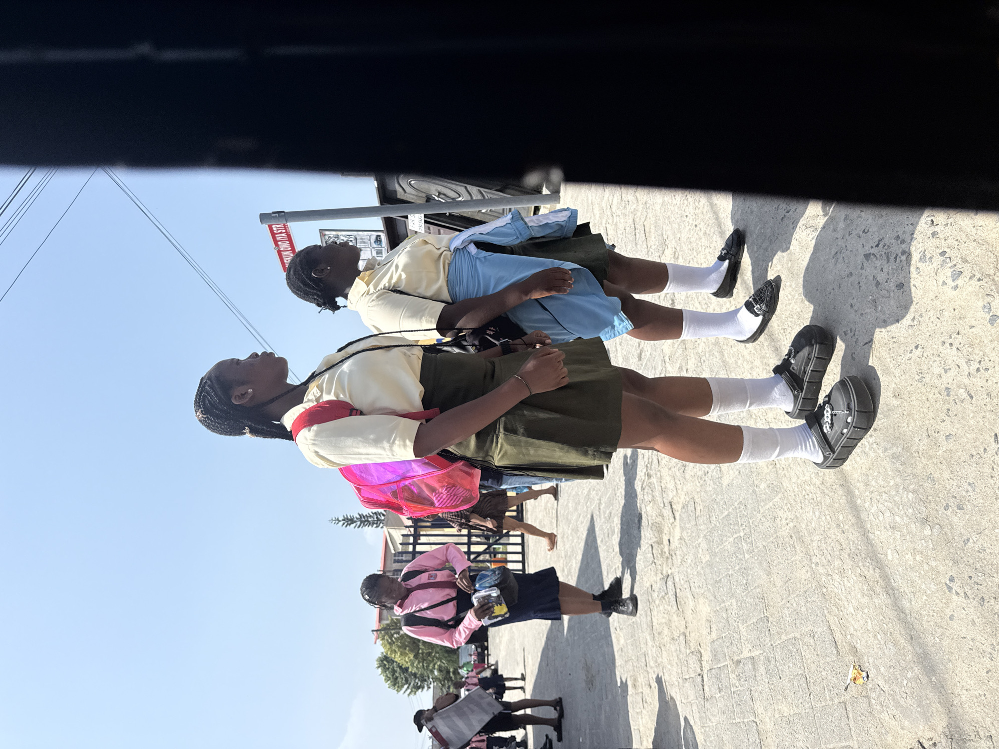 In Ado, Nigeria, two young female students in school uniforms stand outdoors on a paved walkway. The student on the left wears a cream-colored short-sleeved blouse and a dark green pleated skirt, with white socks and black clogs. She carries a bright pink backpack and a dark bag. The student on the right, also in a cream-colored blouse and dark green skirt, has a light blue jacket tied around her waist, white socks, and black loafers. She carries a dark bag on her shoulder. Behind them, a street sign reads "IKUN OWO IYA STR". Other students and a fence are visible in the background, with trees and buildings further in the distance. The scene is illuminated by bright sunlight, casting shadows on the ground.