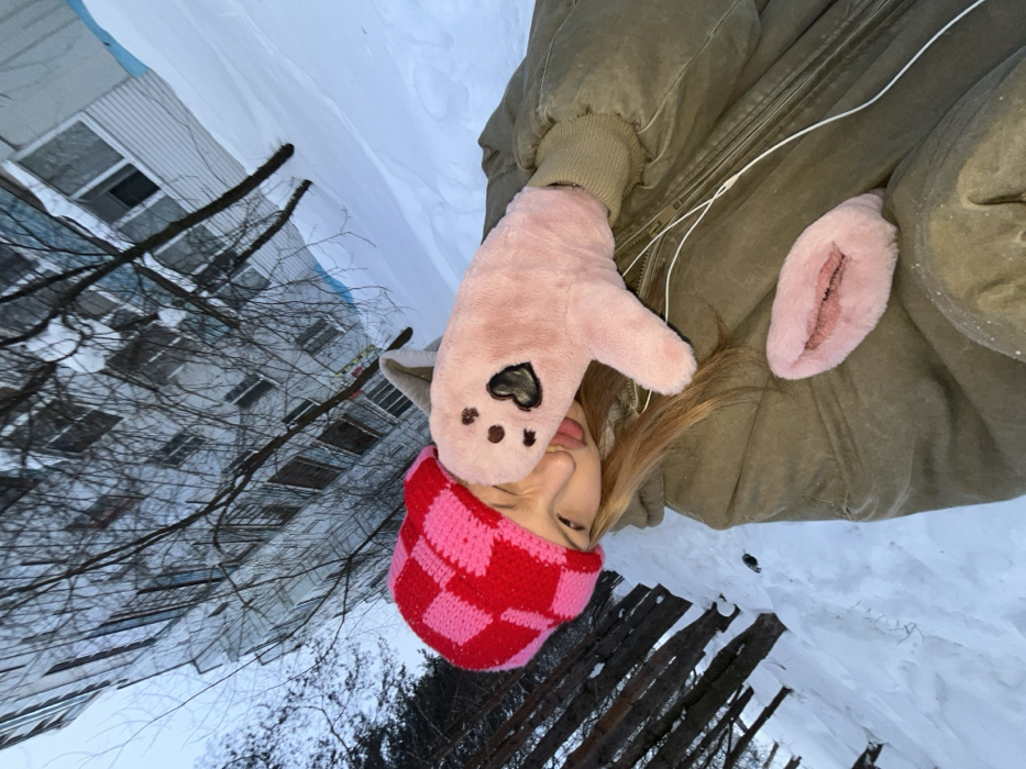 An individual is captured in a selfie in a snowy outdoor environment in Chaikovsky, Russia. The subject, a person with blonde hair, wears a red and pink checkered knitted beanie and an olive-green winter jacket. The person's tongue is extended, touching a light pink, furry mitten on their right hand, which displays dark paw-print-like markings and a heart emblem. A smaller, pink furry object, shaped like lips or a vulva, rests on the jacket below the mitten. White wired earbuds are visible draped across the jacket. The background features extensive snow coverage, bare deciduous trees with dark trunks, and a multi-story, light-colored apartment building with numerous windows under an overcast sky, indicative of winter conditions. The image is taken from a low, upward angle.