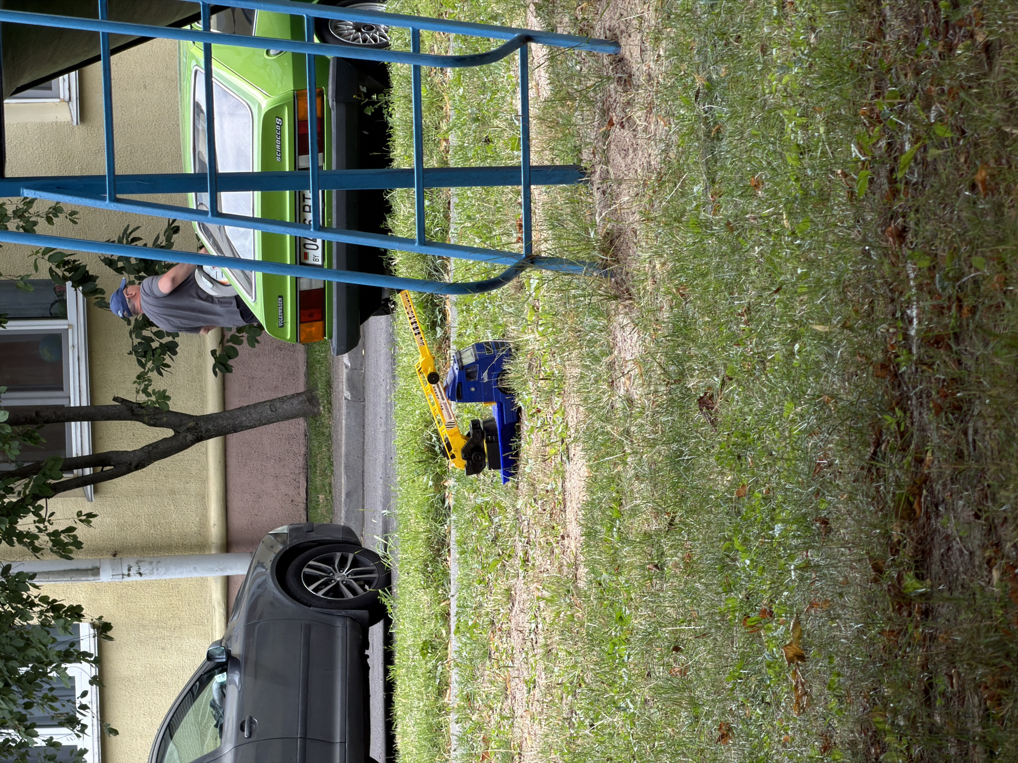 In Minsk, Belarus, on a sidewalk next to a building with a yellow facade, a gray car and a green Volkswagen Scirocco are parked. In front of the cars, a blue toy crane is on the grass. A man wearing a blue cap and a gray t-shirt stands near the green car, holding a white helmet. The scene appears to be outdoors during the daytime.