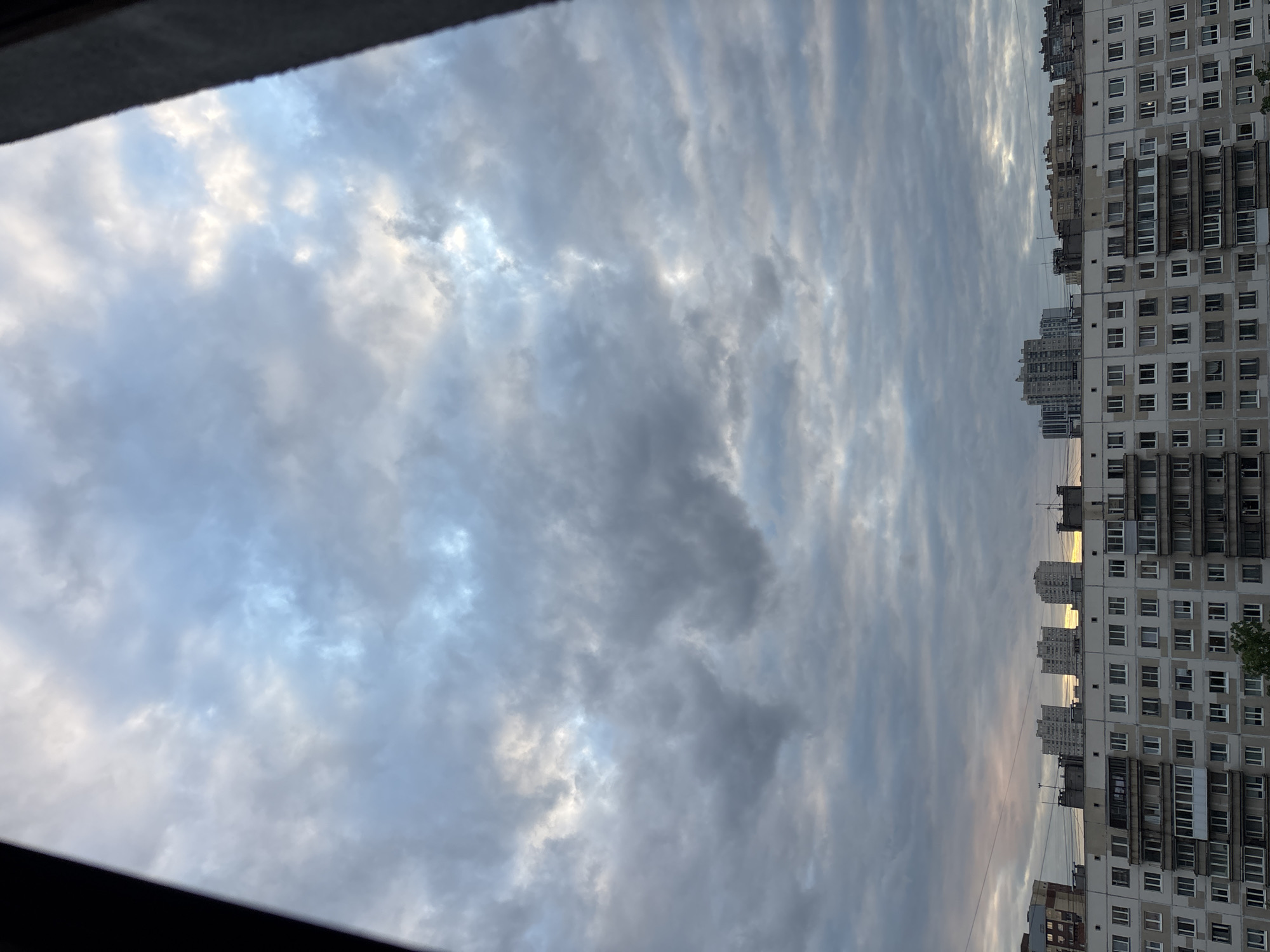 The image captures a view from a window in Vsevolozhsk, Russia. The sky is predominantly filled with altostratus clouds, layered horizontally across the frame. Patches of blue sky are visible amidst the cloud cover. Along the right edge of the image, a dense apartment building is visible, with multiple rows of windows. The building appears to be typical of Soviet-era construction. The overall impression is of an overcast day in a densely populated urban area.