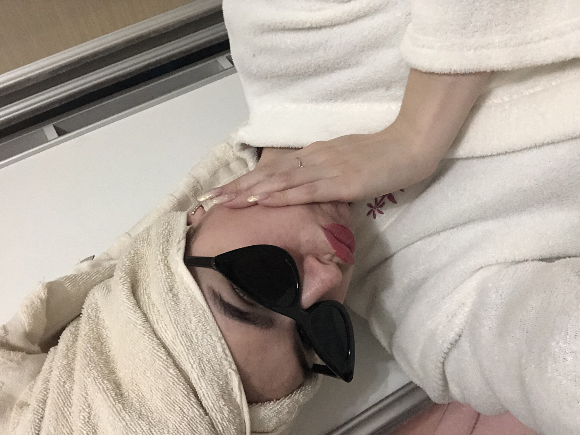 An individual wearing black sunglasses and a towel wrapped around their head lies on a white surface. The person is also wearing a white bathrobe, and a hand with a ring is placed on their face. The location cannot be determined based on the provided image.