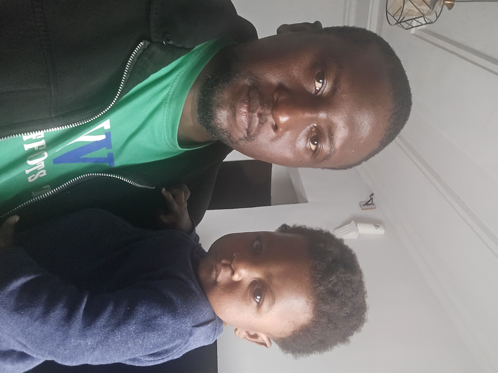 A medium shot captures a Black man holding a young Black child. The man is in the foreground, facing forward with a neutral expression. He has dark skin, a shaved head, and a short beard. He is wearing a green t-shirt with a graphic design and a black jacket zipped up. The child is nestled in the man's arms, facing slightly to the right. The child has dark, curly hair and is wearing a blue long-sleeved shirt. The background is out of focus, but appears to be an indoor setting with a white ceiling and a light fixture on the left. The lighting suggests a well-lit room. The image does not contain any information or visual cues to determine its location in Jos, Nigeria.