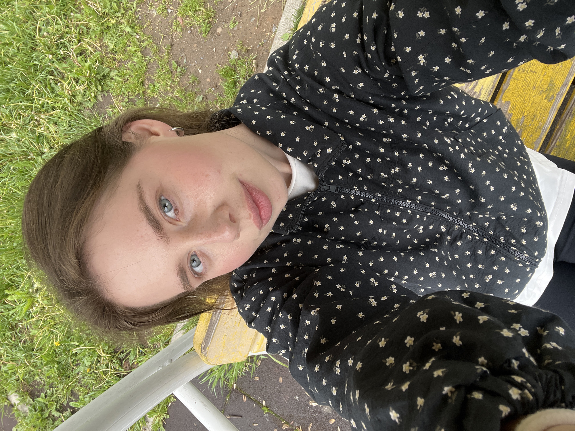 The image is an upward-angled selfie of a young individual, likely female, captured outdoors in what appears to be a park or public green space in Chaikovsky, Russia. The person has fair skin, light brown hair, and striking blue eyes, looking directly at the camera with a neutral to slightly thoughtful expression. Their head is tilted, giving the shot a casual feel.
They are wearing a black bomber jacket adorned with a pattern of small white and yellow floral designs, with a white top visible underneath. A silver hoop earring is visible in their left ear. The background features patches of vibrant green grass and some bare earth. A yellow wooden bench or structure is visible behind the person, and a white railing or fence runs diagonally across the foreground. The ground appears somewhat damp, suggesting recent precipitation or morning dew. The natural, diffused lighting indicates it is daytime, possibly under an overcast sky. There is no visible text in the image.