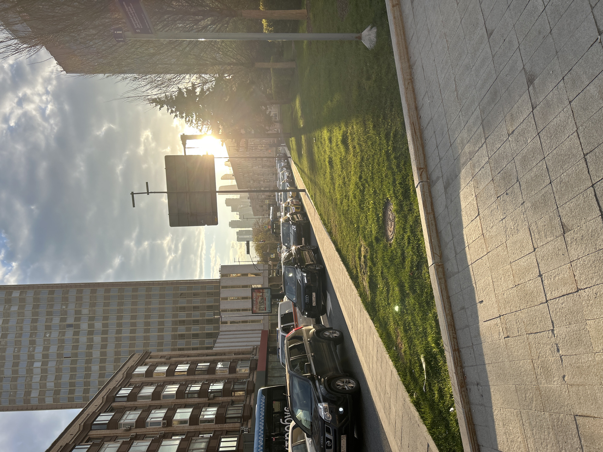 The image captures an urban street scene during the late afternoon or early evening, characterized by warm, low-angle sunlight casting long shadows.
On the left side of the frame, there's a multi-story building with a modern, gridded facade, reflecting the sky. Below it, an older, brick building with numerous windows is visible. The street runs horizontally through the middle ground, lined with a queue of parked or slow-moving cars, including several SUVs (a black Audi with license plate "H208AM 77" and a black Nissan X-Trail with a partial plate ending in "150") and a black bus with "SAYBUS" written on its side. On the right, a wide grassy median with trees separates the road from a paved sidewalk, made of large square pavers, that extends into the foreground. The grass is vibrantly lit by the sun, while parts of the sidewalk are shaded.
The sun is prominently visible on the horizon, shining brightly through scattered clouds, creating a strong lens flare and illuminating the scene with a golden glow. A pole with a street sign, including an "i" symbol for information and Cyrillic text ("Информация о гостиничных и других организациях" - "Information about hotel and other organizations"), stands near the top left. Another large, blank sign is visible in the foreground. In the distance, a city skyline with more buildings can be seen. A poster on one of the buildings reads "ТОРГОВЫЙ..." (likely part of "Торговый Центр" - "Shopping Center"). The license plate codes '77' and '150' suggest a location in or around Moscow, Russia.