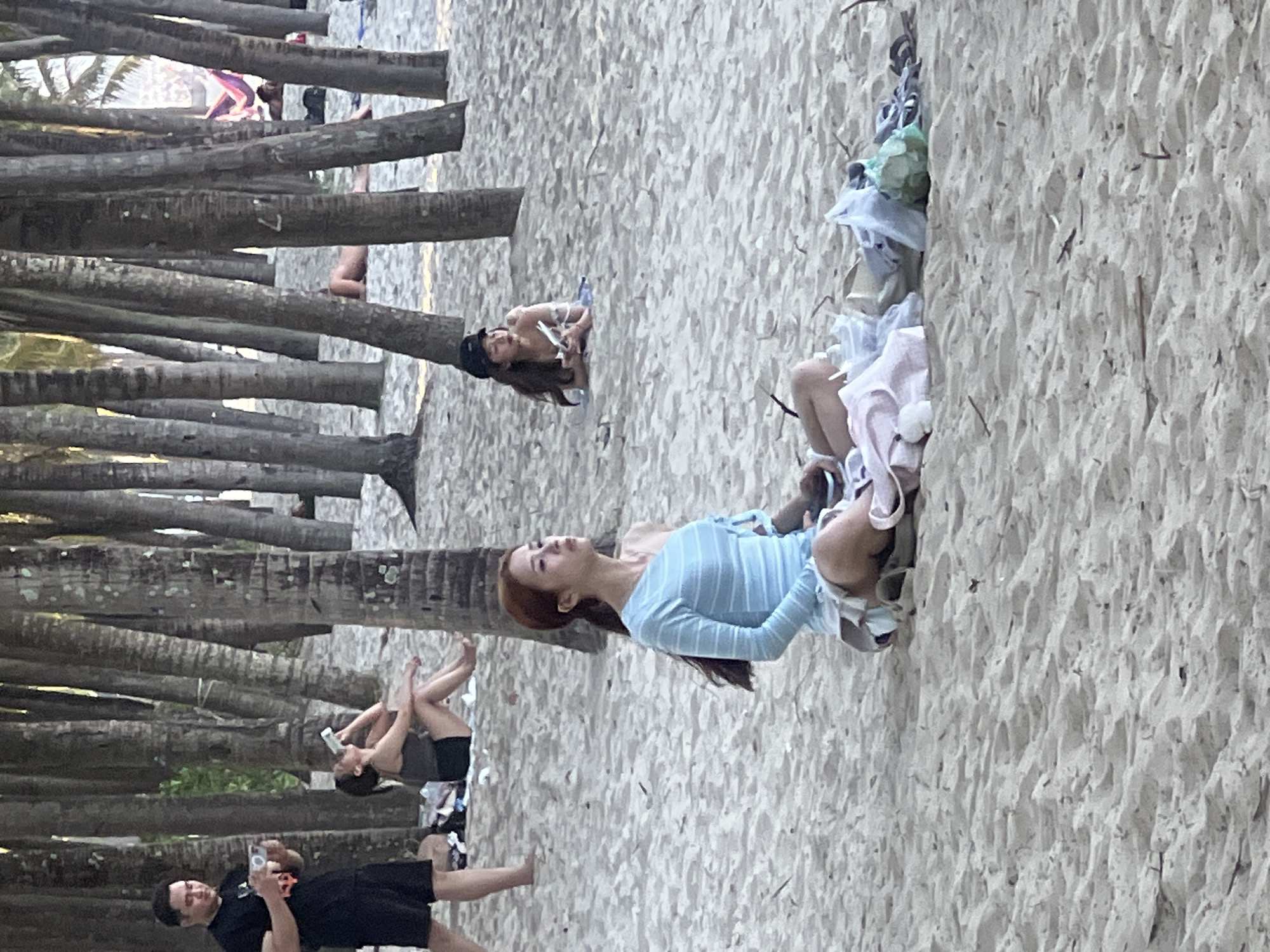 This image depicts a crowded beach scene in Da Nang, Vietnam, characterized by a dense line of palm trees running perpendicular to the shoreline. In the foreground, a woman with reddish-brown hair is sitting cross-legged on the sand, wearing a light blue, off-the-shoulder striped top and a white skirt. She is looking to her right, with her hands resting on a pile of clothing and plastic bags.
Further back, on the left side of the image, a man in a black t-shirt and shorts is standing and holding a mobile phone. To his right, a woman in a dark tank top and shorts is seated, drinking from a white bottle. In the middle ground, another woman is lying on a blue towel, wearing a black cap and holding a phone. In the background, several people are visible, some lying on the sand, and a hammock is suspended between two palm trees. The beach is covered in sand, with scattered debris from beachgoers. The lighting suggests it is daytime, possibly late afternoon.