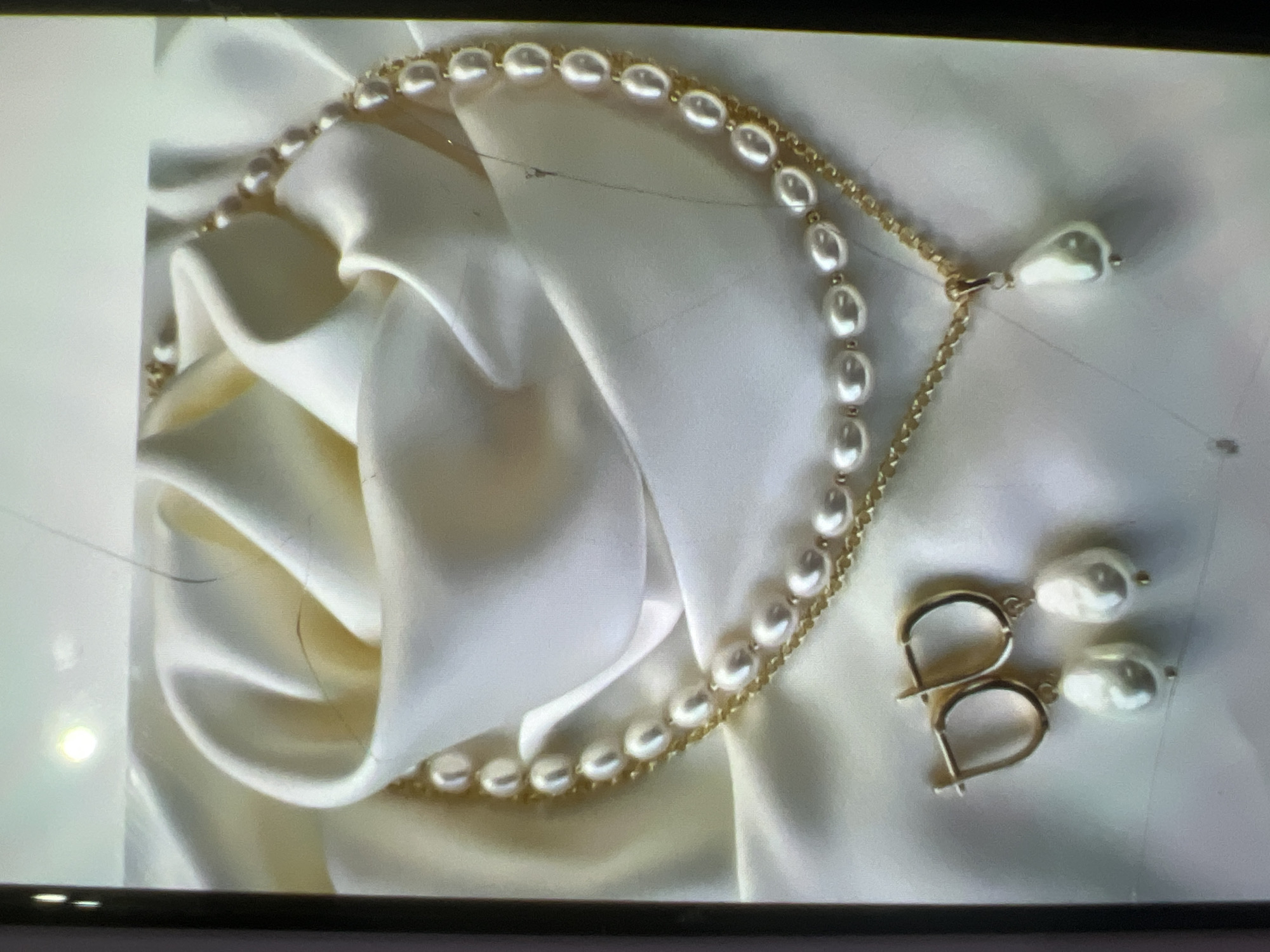 The image shows a close-up view of a pearl necklace and earrings set on white satin fabric. The necklace is composed of two strands of pearls. The upper strand is a simple pearl necklace. The lower strand is a gold chain with a single pendant pearl in a teardrop shape. To the left of the necklace are a pair of gold hoop earrings, each with a single pearl hanging from it. The fabric appears to be draped in a way that resembles a rose or other floral form. The lighting is soft and diffused. There are no people or any elements indicating a specific location in Kara-Dıykan, Kyrgyzstan.