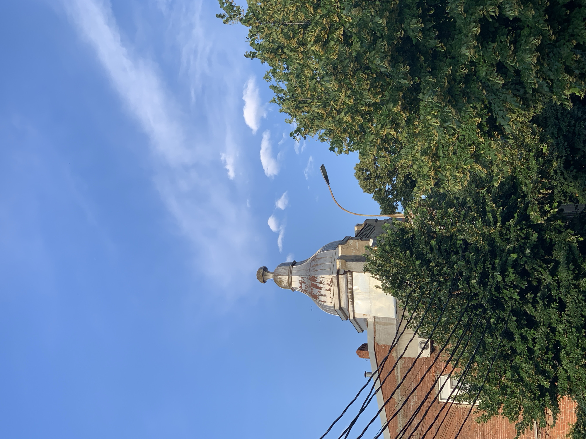 A low-angle, outdoor, daytime shot captures a clear blue sky with scattered white clouds. In the foreground, a cluster of dark electrical wires runs diagonally from the bottom left across the frame. Behind the wires, a brick building with white trim and an air conditioning unit is visible. To the right of the brick building, a tall green tree with small yellowish blossoms occupies a significant portion of the right side of the image. Peeking above the trees and behind the wires is the top of a domed, weathered building with a metallic, rusted roof. A street lamp on a curved pole stands to the left of the domed building.
