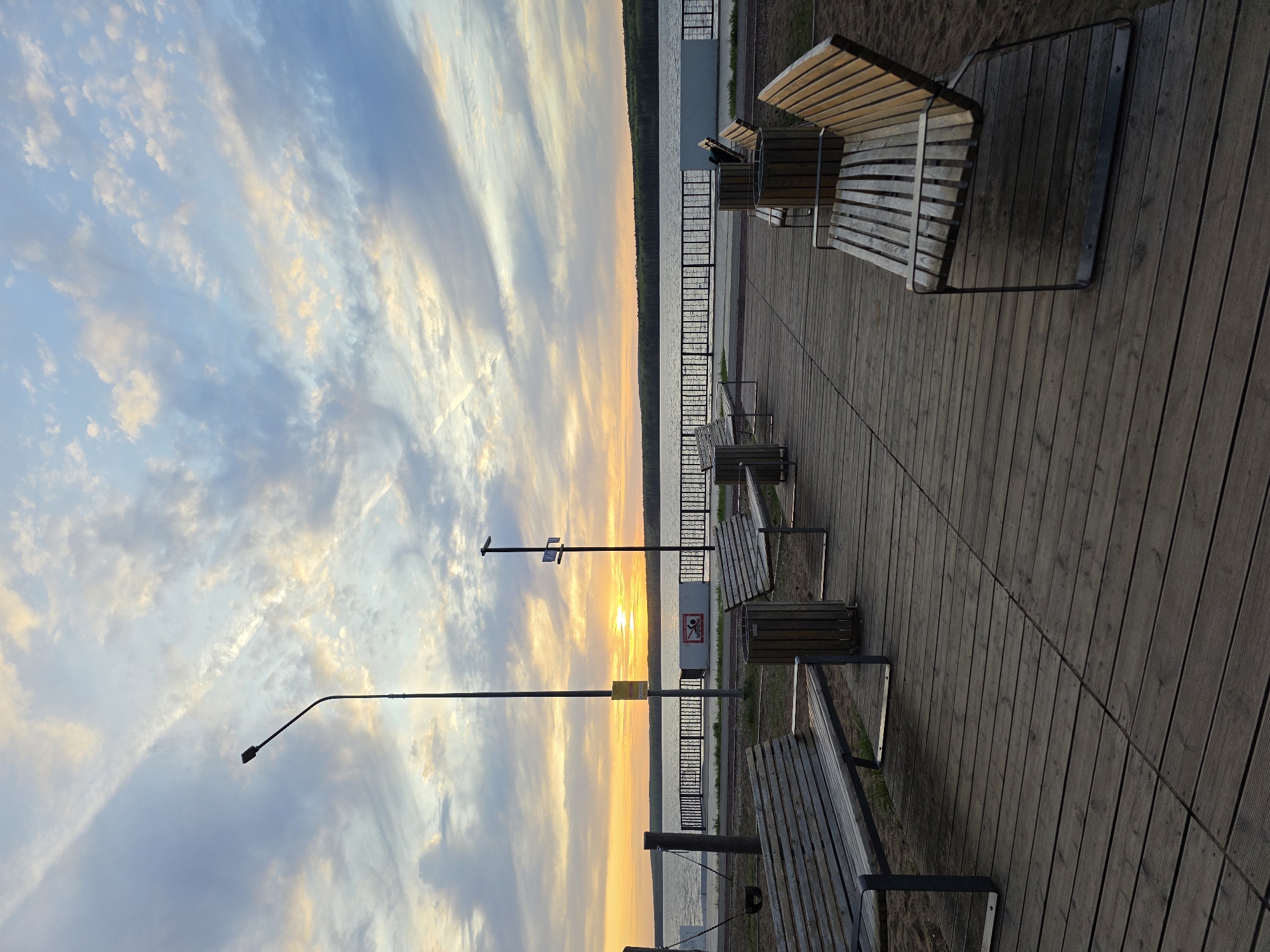 The image shows a row of wooden benches on a boardwalk overlooking a body of water, likely a lake or river. The sky is filled with streaks of clouds illuminated by the setting sun, which is visible on the horizon. There are street lights along the boardwalk. A black metal fence separates the boardwalk from the water. The image appears to be taken in the city of Chaikovsky, Russia, possibly near a popular recreation area or waterfront.