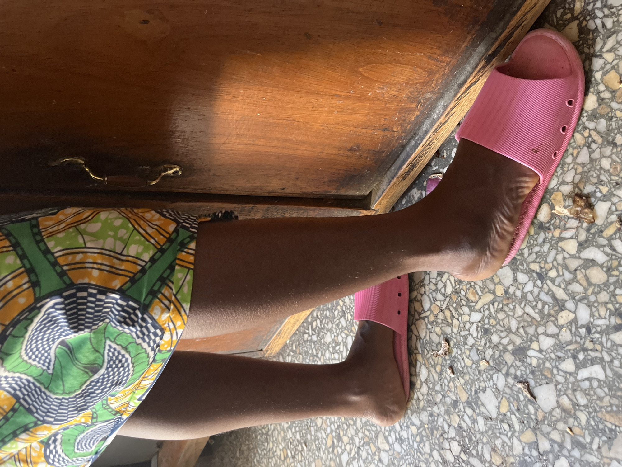 The image shows the lower legs and feet of a person wearing a patterned dress in shades of green, yellow, and black, and pink plastic slides. The person is positioned next to a wooden object, possibly a desk or cabinet, with a drawer pull visible. The floor is a mottled grey and white tile. There is no contextual information in the image to confirm the location in the city of Jos, Nigeria.