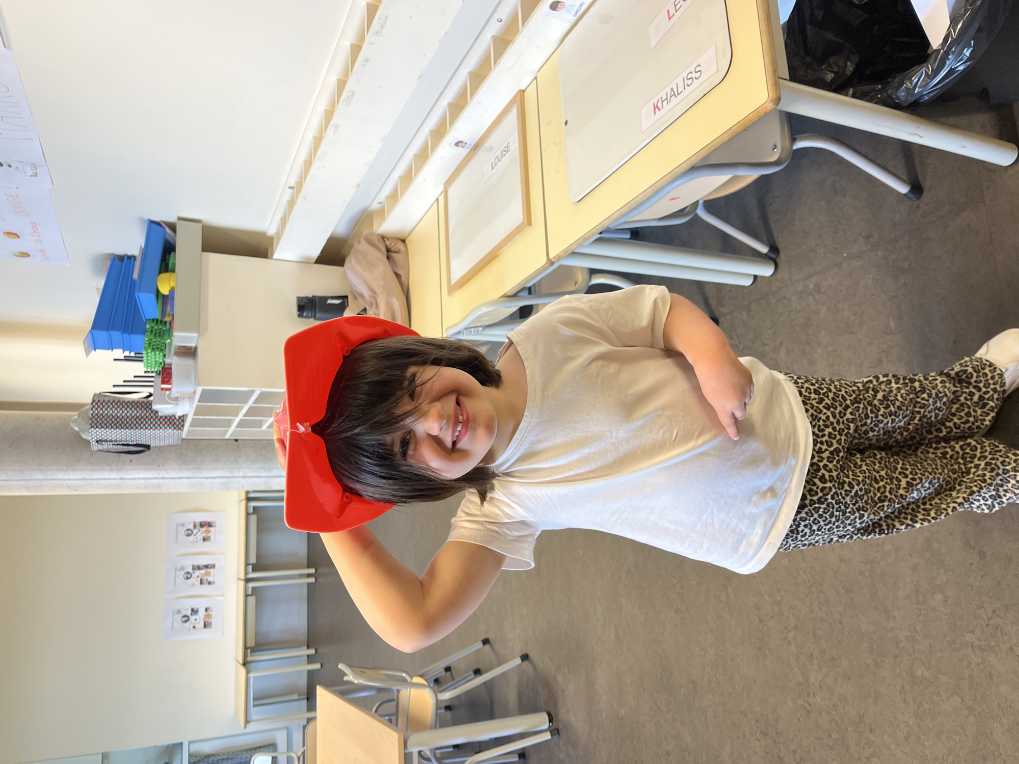 The image captures a smiling young child in what appears to be a classroom setting in La Fouillouse, France. The child has dark hair, is wearing a white short-sleeved t-shirt and leopard-print trousers, and holds a bright red hat on their head. They are looking directly at the camera with a happy, playful expression.
The background shows typical classroom furniture and elements. Several light-colored desks and chairs are visible, with two desks prominently featuring name tags: "LOUISE" and "KHALISS." A partial name, "LE," is also visible on another tag. A black trash bag is positioned next to one of the desks. Along the walls, there are storage units with shelves and cubbies, holding items like blue stacking trays and green interlocking gears. Educational papers are affixed to the walls, one partially displaying the word "ORANGE!". The floor is a solid, neutral grey. The lighting is bright and even, characteristic of an indoor daytime environment.