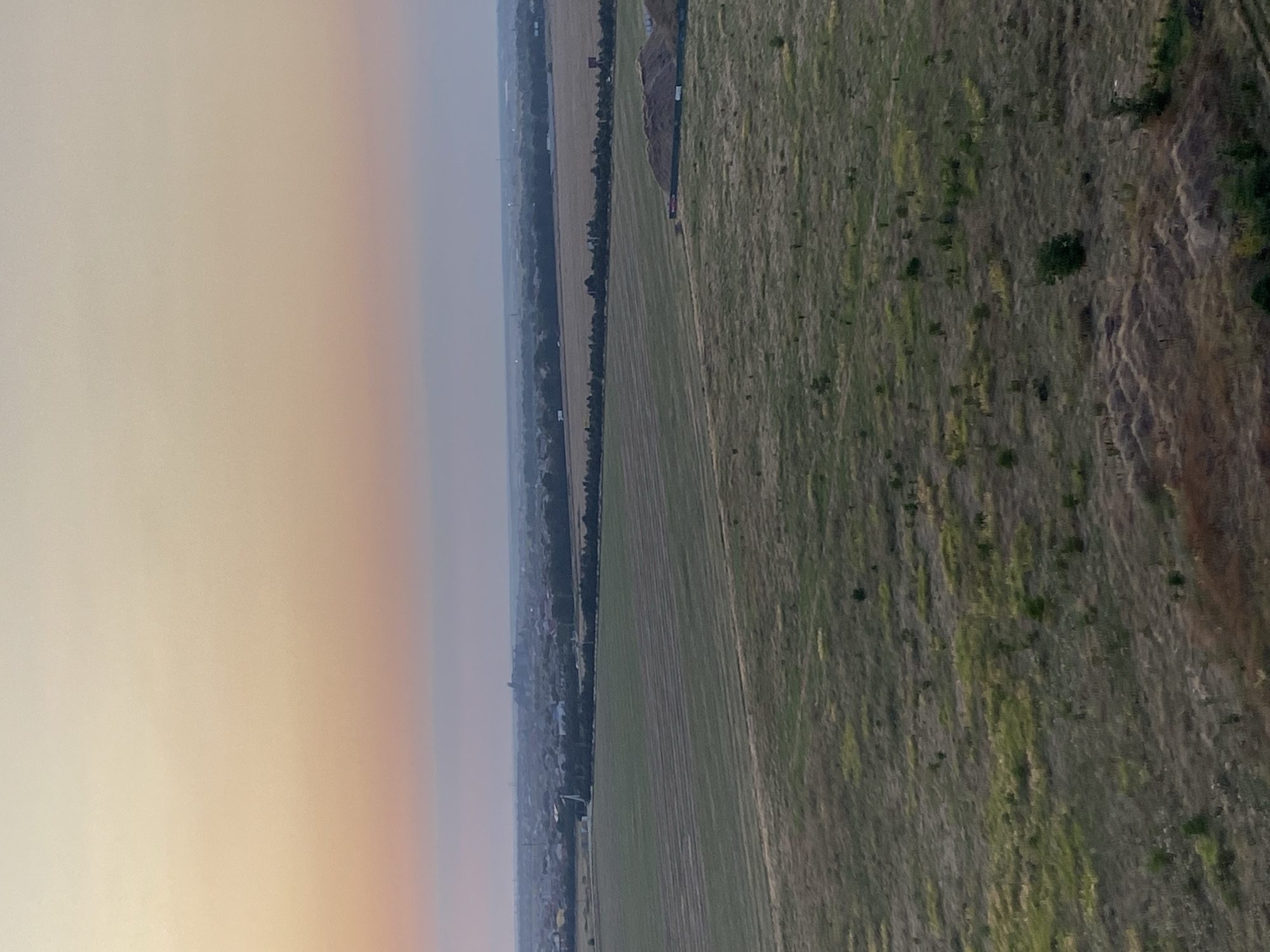 The image captures a wide aerial view of a landscape at dusk, likely on the outskirts of Chornomorsk, Ukraine. The sky exhibits a gradient of warm colors, transitioning from pale yellow at the top to soft pink and then a muted blue closer to the horizon. Below the sky, a sprawling panorama of agricultural fields dominates the scene. The fields are characterized by varying textures and colors, with some appearing as neatly tilled plots with striped patterns, while others are covered in wilder, drier grasses and scattered vegetation. In the mid-ground, a treeline separates the agricultural fields from a settlement, which is partially visible. The settlement comprises numerous buildings with dark roofs, indicative of a residential area or small town. Further in the distance, the landscape appears hazy, with faint structures and possibly industrial elements visible on the horizon. On the right side of the image, a large mound of dirt or aggregate material is present, suggesting construction or excavation activity. The overall impression is one of rural expanse meeting encroaching development under a serene twilight sky. There are no people or animals visible in the image.