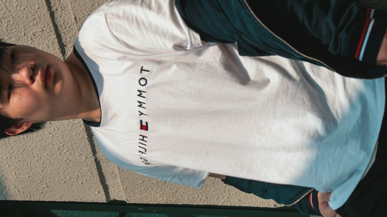 In Oreshki, Russia, a young person stands against a textured, light-colored wall. They are wearing a white t-shirt with the Tommy Hilfiger logo printed on it. The t-shirt has a dark trim around the neckline. Over the t-shirt, they are wearing a dark jacket, which is partially unzipped, revealing the white t-shirt. The jacket also has stripes on the sleeves. The lighting suggests it is daytime, with sunlight casting shadows on the wall and the person's clothing. The background is plain, with the textured wall being the dominant feature.
