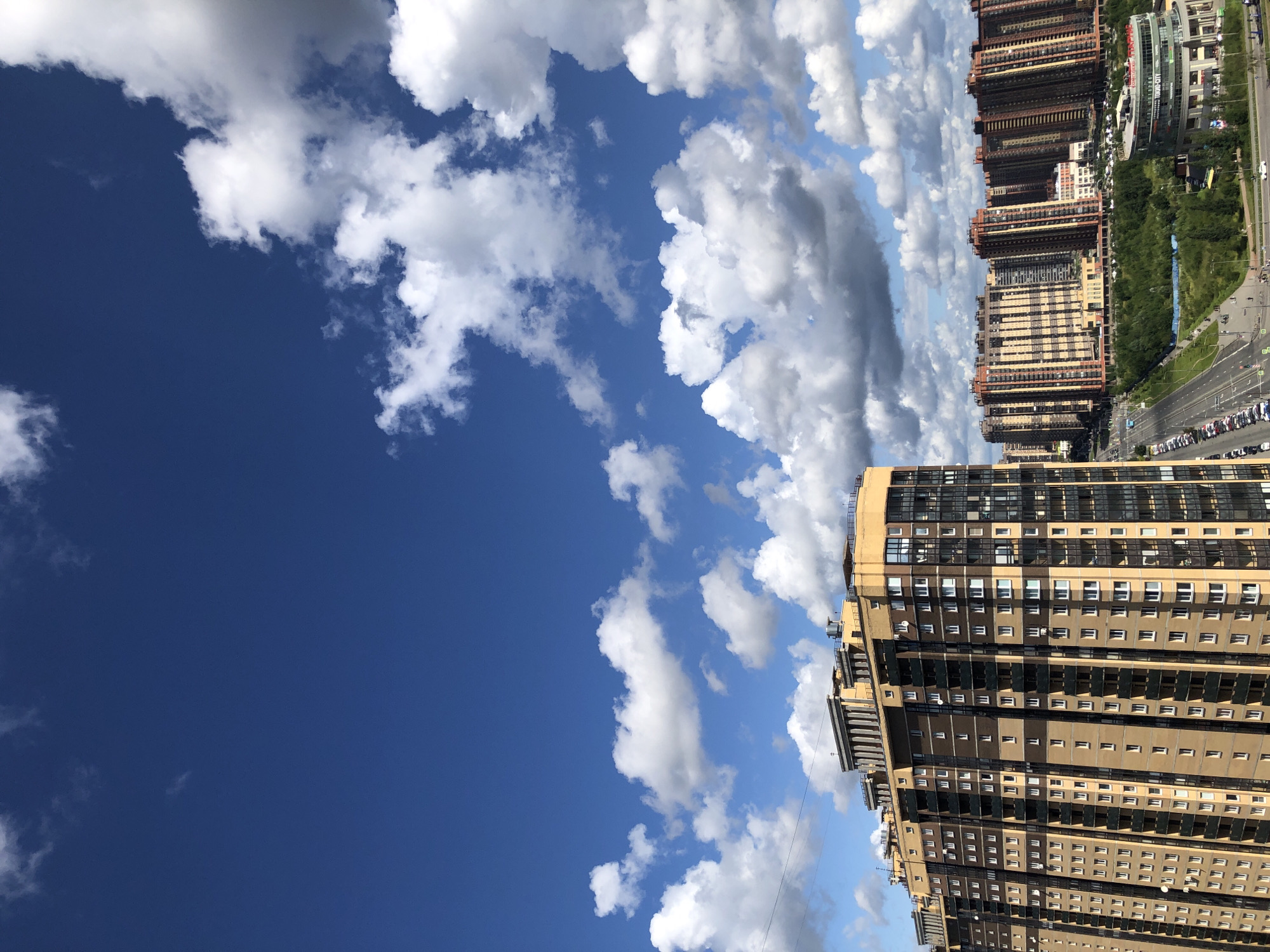 This is a high-angle shot of a modern cityscape under a bright blue sky dotted with fluffy white clouds. The foreground features a tall, multi-story residential building with a distinctive facade of beige and dark brown sections, punctuated by numerous windows and balconies. To the right of this building, a series of similar, densely packed residential towers stretch into the distance, exhibiting a range of warm color palettes. A wide, multi-lane road runs through the middle ground, lined with parked cars and bordered by lush green trees and a curved, modern building with visible signage. The scene suggests an urban development with a focus on high-rise living.