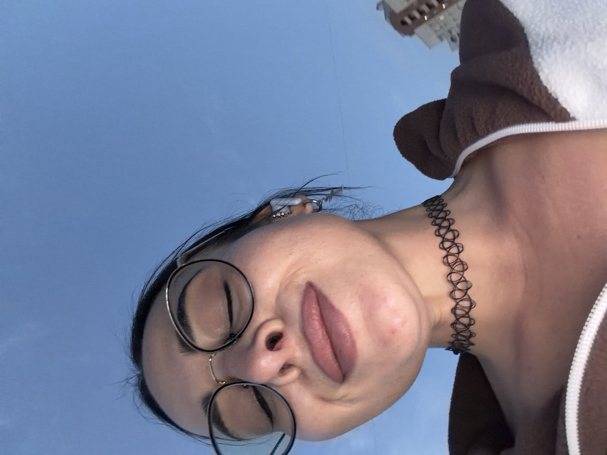 The image is a low-angle, close-up portrait of a young person, likely female, captured from below looking upwards against a clear blue sky. The subject's head is tilted, and their gaze is directed downwards towards the camera. They wear dark, round-framed eyeglasses and have a small hoop piercing in their nose. A silver earring with multiple elements is visible on their right ear. Their lips are painted a muted pinkish-brown and are slightly parted in a pout-like expression. Around their neck, they wear a black, stretchable choker necklace with an intricate, open-work design. The person is dressed in a cozy-looking garment, predominantly brown and white, which appears to be a hooded jacket or onesie, with a white zipper visible on the right side.
The background consists primarily of a bright, cloudless blue sky, indicating daytime. In the upper right corner, a portion of a light-colored, multi-story building with windows is faintly visible, suggesting an urban or semi-urban setting. Given the context of Saint Petersburg, Russia, this outdoor shot captures a moment of casual posing. There is no visible text in the image. The overall mood is casual and direct, with a focus on the subject's face and accessories.